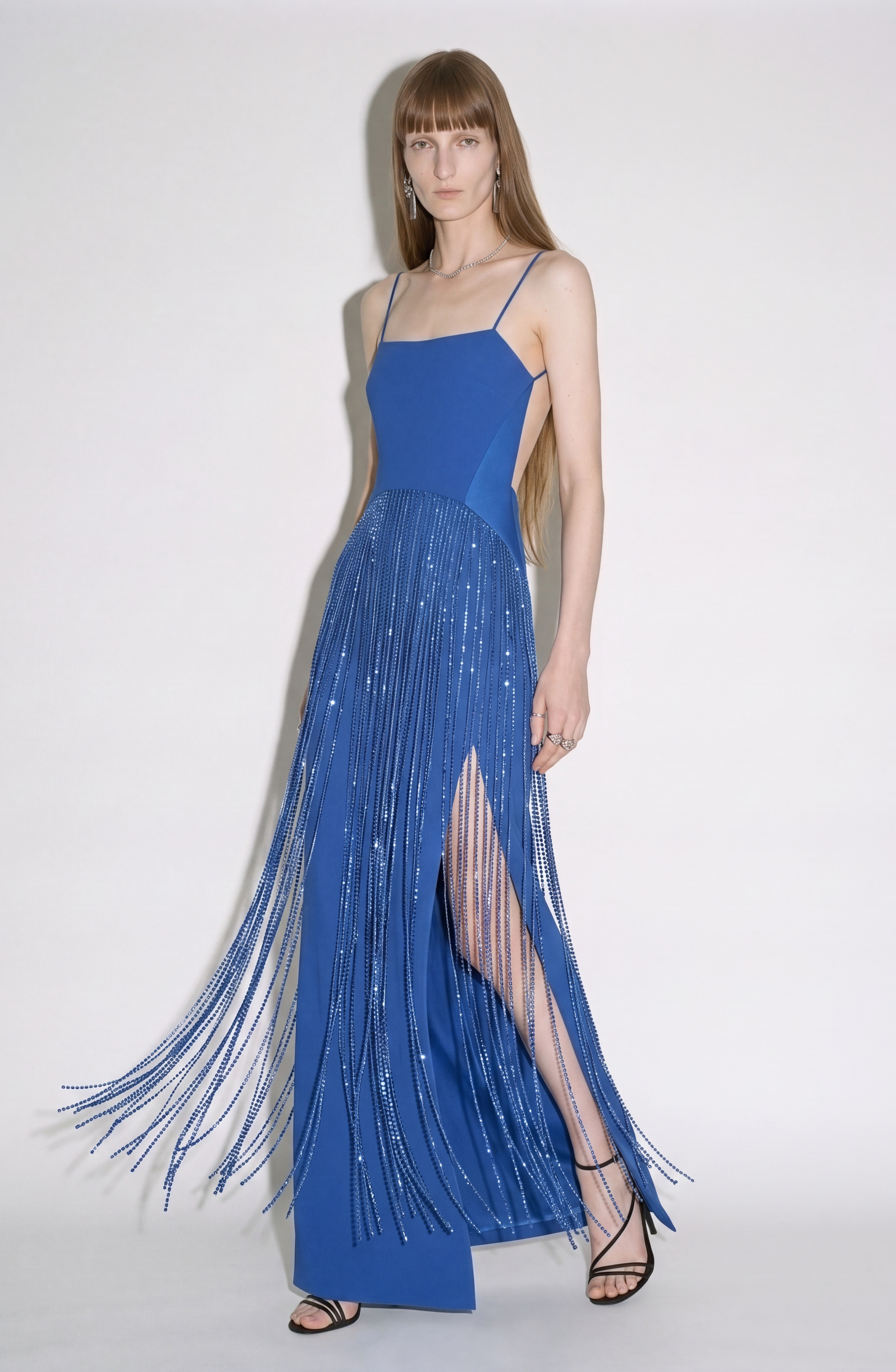 A woman stands against a plain white background wearing the LOULOU Cobalt Crystal Fringe Maxi Dress. Crafted from a luxurious crepe blend, the dress features a crystal-fringed skirt and a thigh-high slit, which she pairs elegantly with black strappy heels.