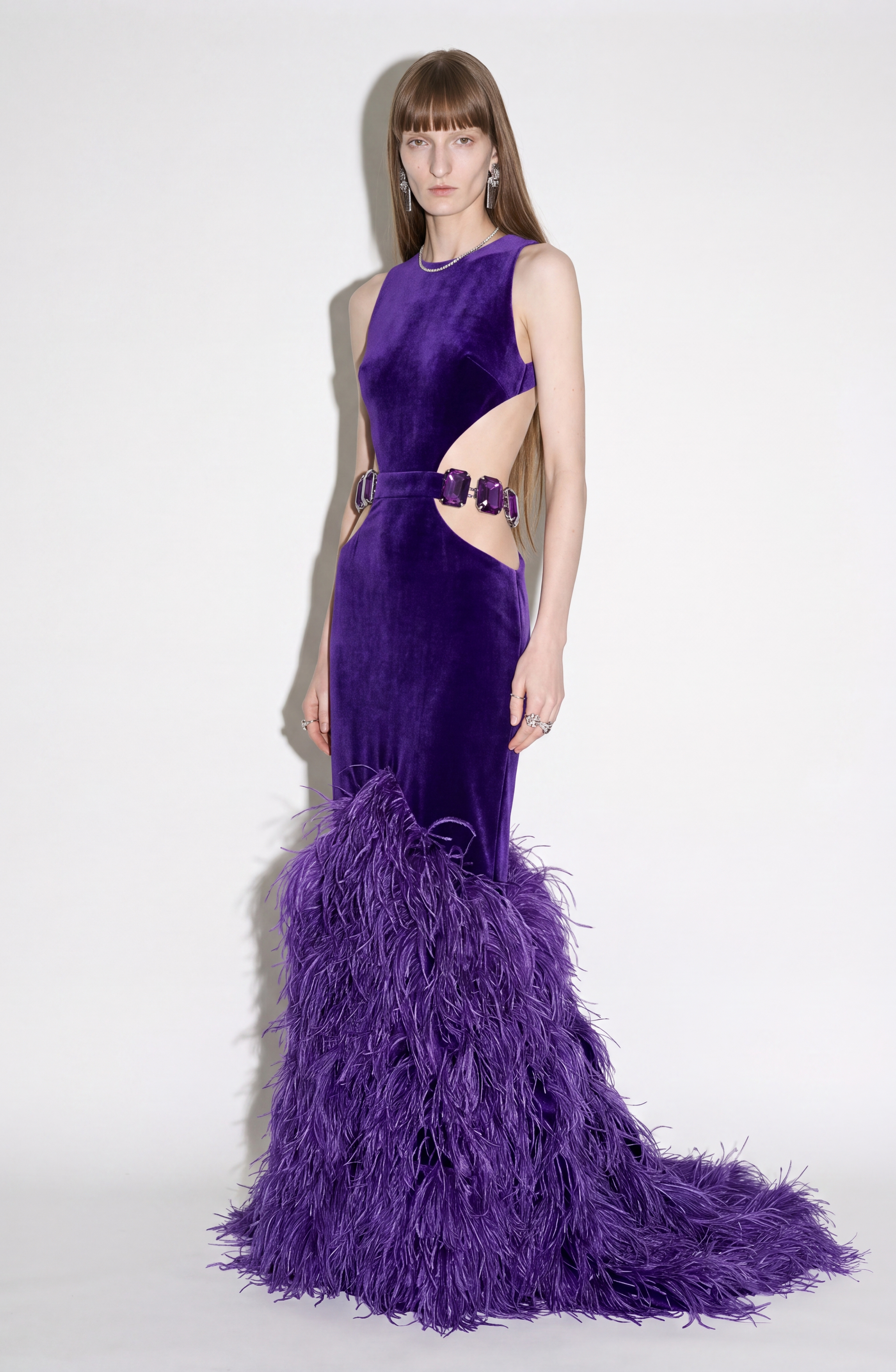 A model wears the LOULOU Velvet Cutout Feather Gown—a sleeveless, purple velvet evening dress adorned with crystal buckles and a dramatic feather-trimmed skirt with side cutouts—posing against a plain white background.