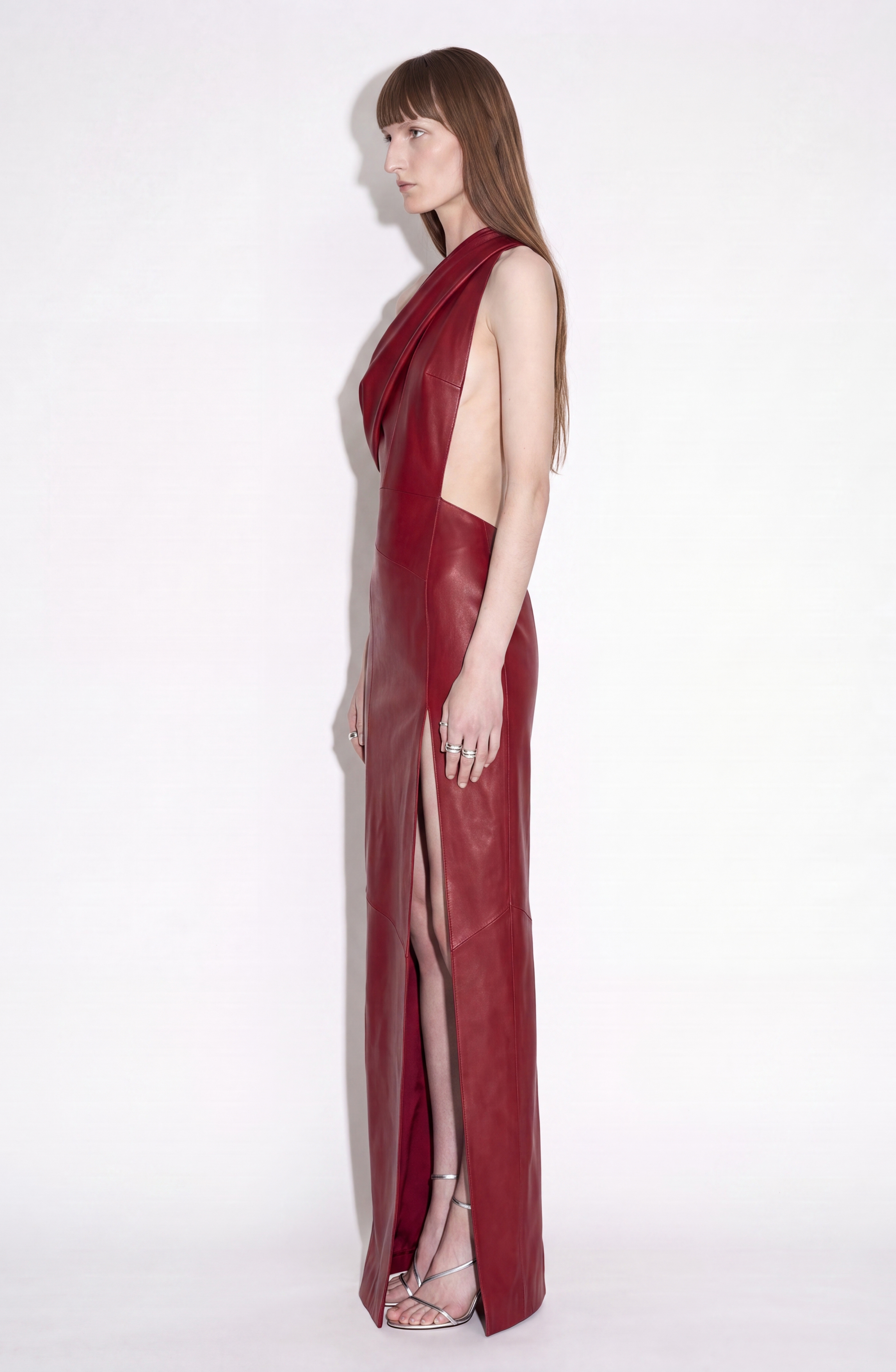 A person stands in profile wearing the LOULOU One-Shoulder Leather Maxi Dress in a rich oxblood hue, featuring a high slit and open sides. The striking ensemble is paired with silver strappy heels, all set against a plain white background.