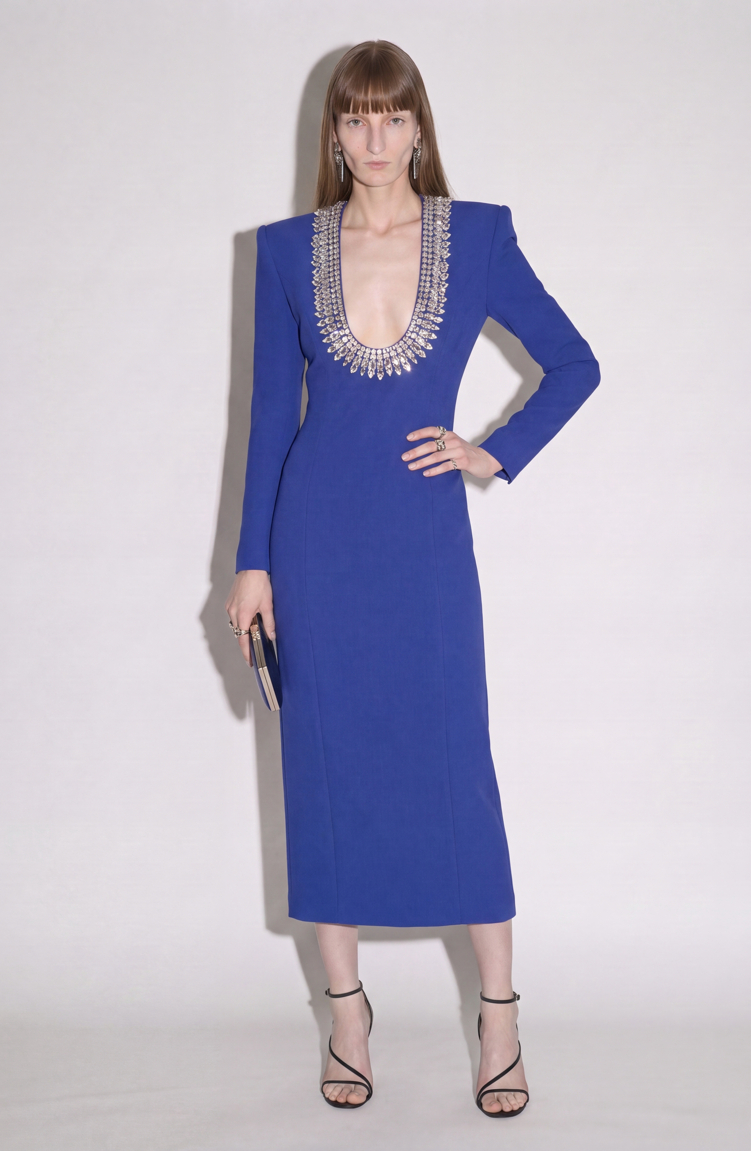 A woman stands against a plain white background wearing the LOULOU Crystal U-Neck Midi Dress in royal blue, featuring long sleeves, padded shoulders, and a deep, crystal-embellished neckline. She completes the look with a clutch and strappy heels.