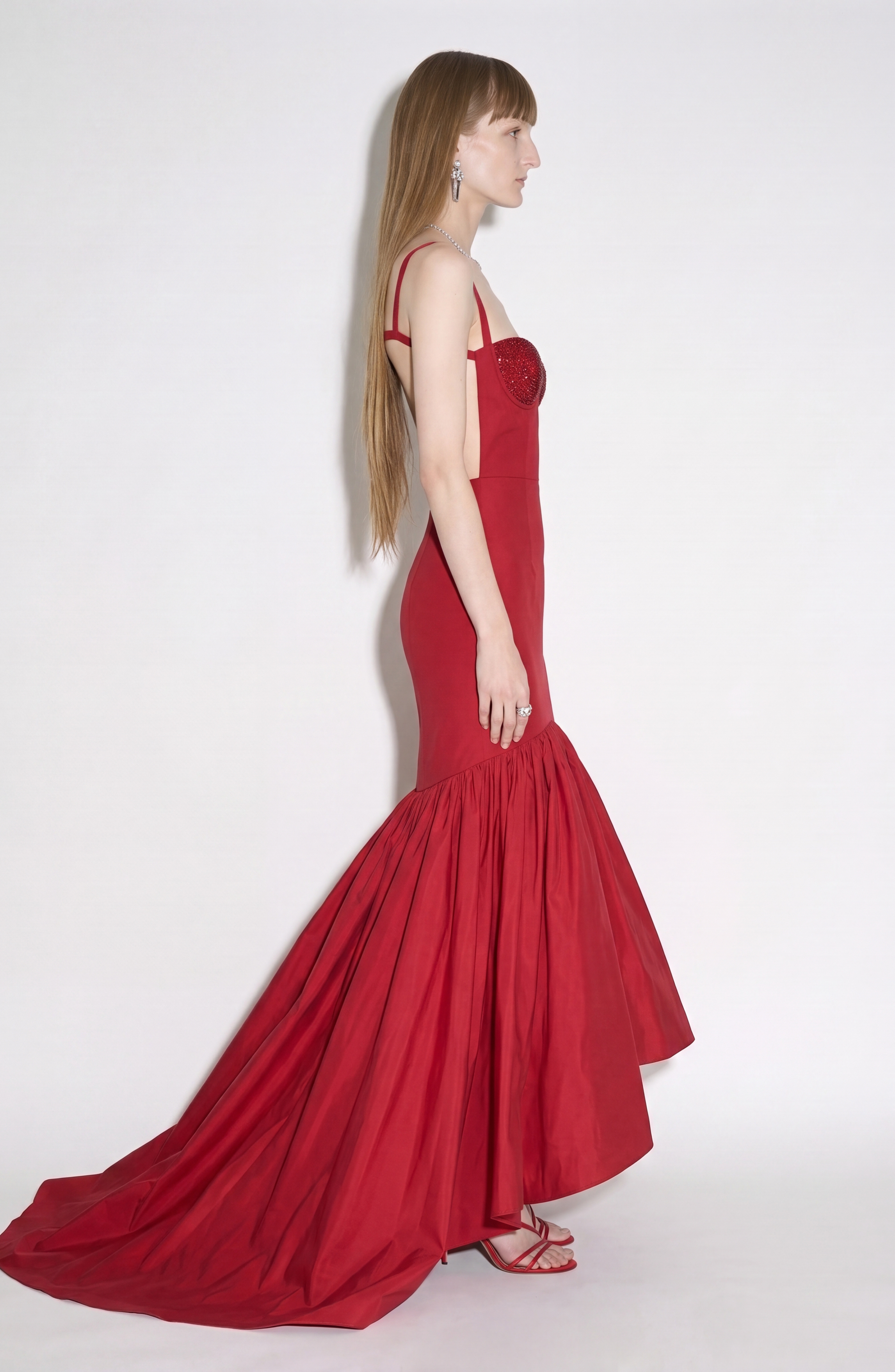 A woman in profile showcases the LOULOU Crystal-Embellished Bustier Gown, featuring a ruby red hue with a sparkling crystal-adorned bodice and a dramatic flared mermaid skirt and train, against a plain white background. She has long, straight hair and accessorizes with red strappy heels and drop earrings.