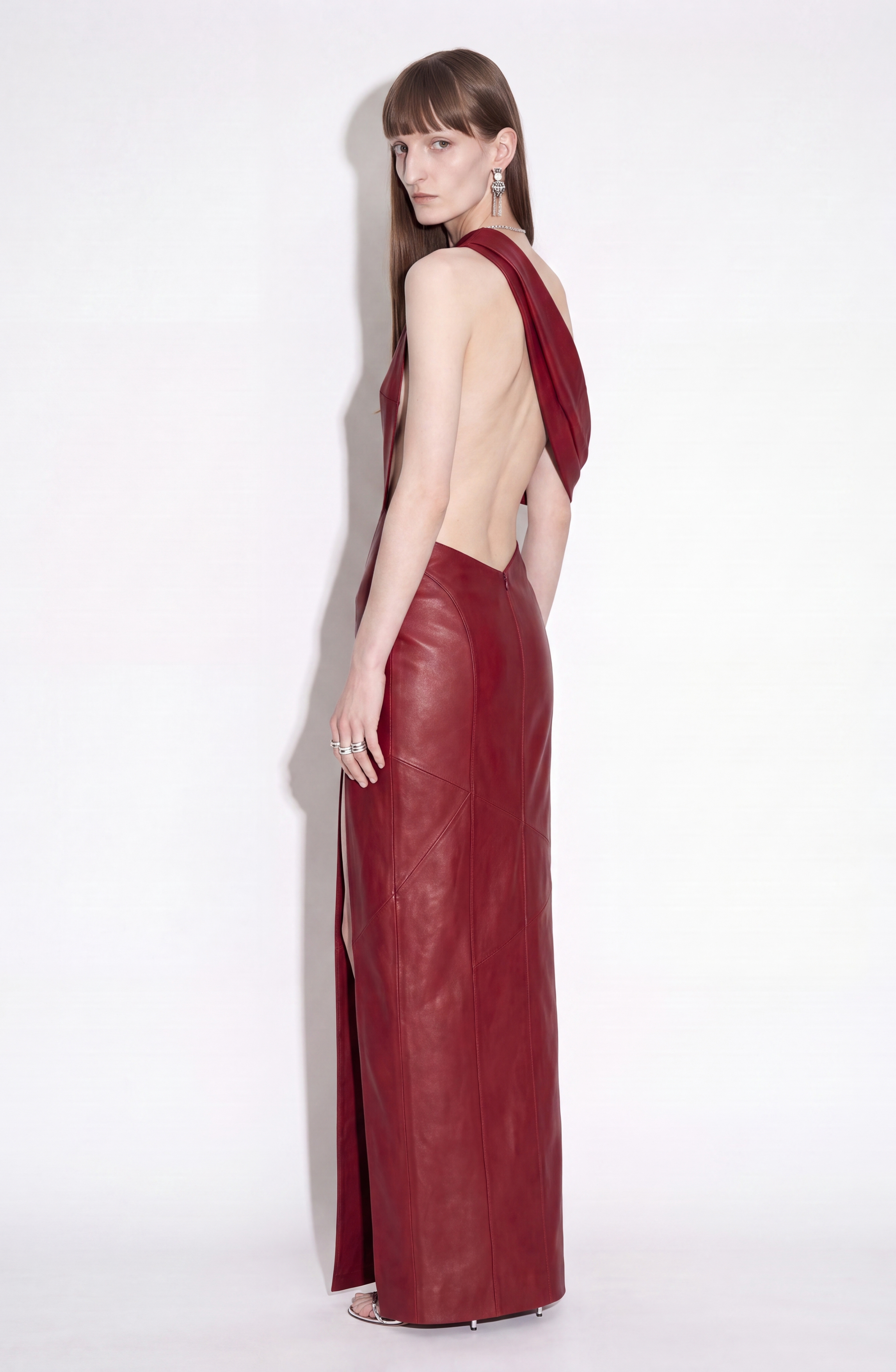 A person stands sideways against a plain white background wearing the LOULOU One-Shoulder Leather Maxi Dress in oxblood, styled with silver earrings and rings.