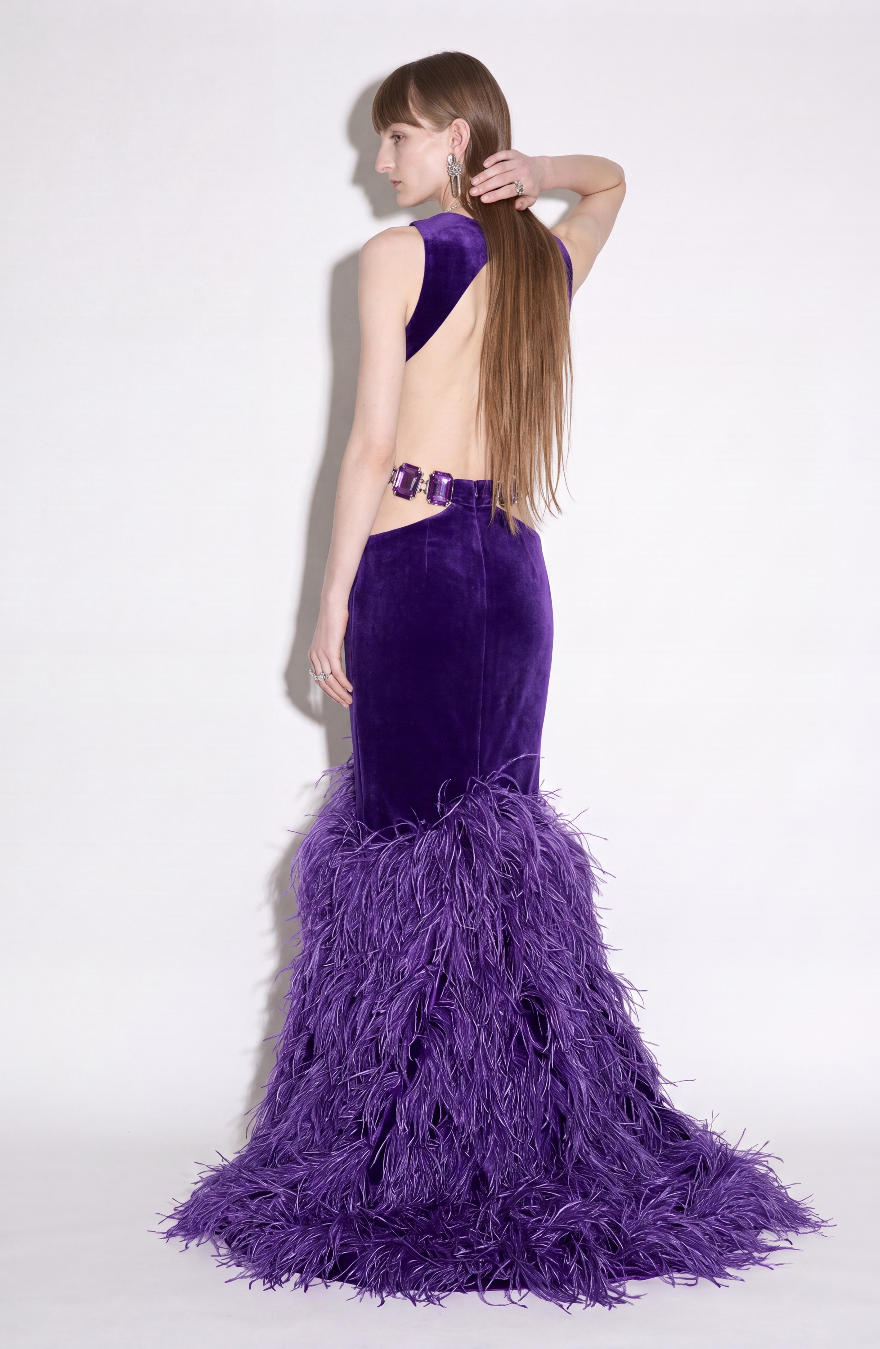 A woman models the LOULOU Velvet Cutout Feather Gown—an elegant, floor-length purple velvet evening dress featuring a backless design, feather-trimmed skirt, and sparkling side crystal buckles—as she stands against a plain white background, facing away and looking to the left.