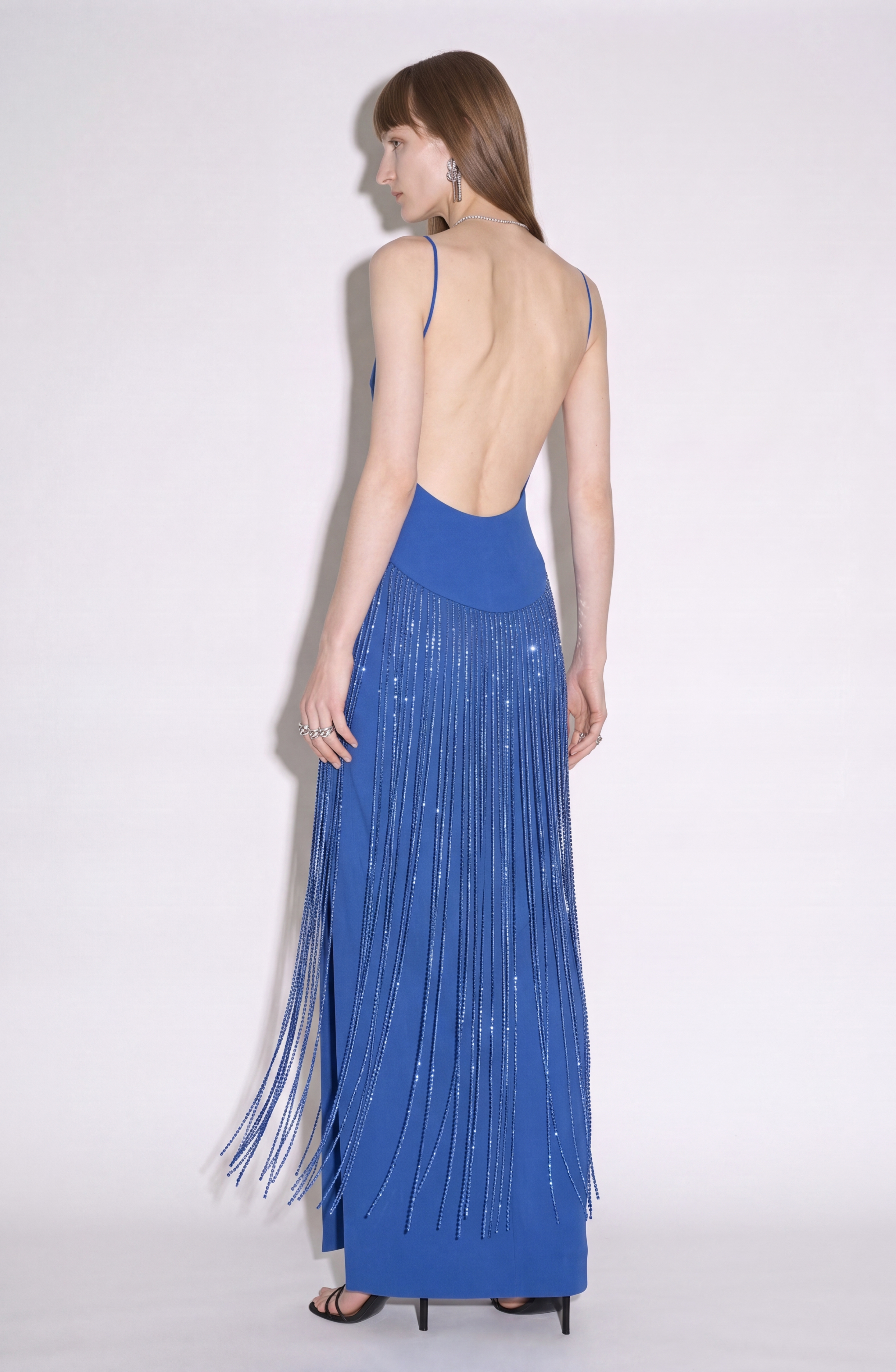 A woman models the LOULOU Cobalt Crystal Fringe Maxi Dress, crafted from a luxurious crepe blend in vibrant cobalt blue. The dress features slender straps and a striking crystal-fringed skirt cascading from the waist. Posed against a plain white backdrop, she faces away from the camera to highlight the gown’s elegant backless design.