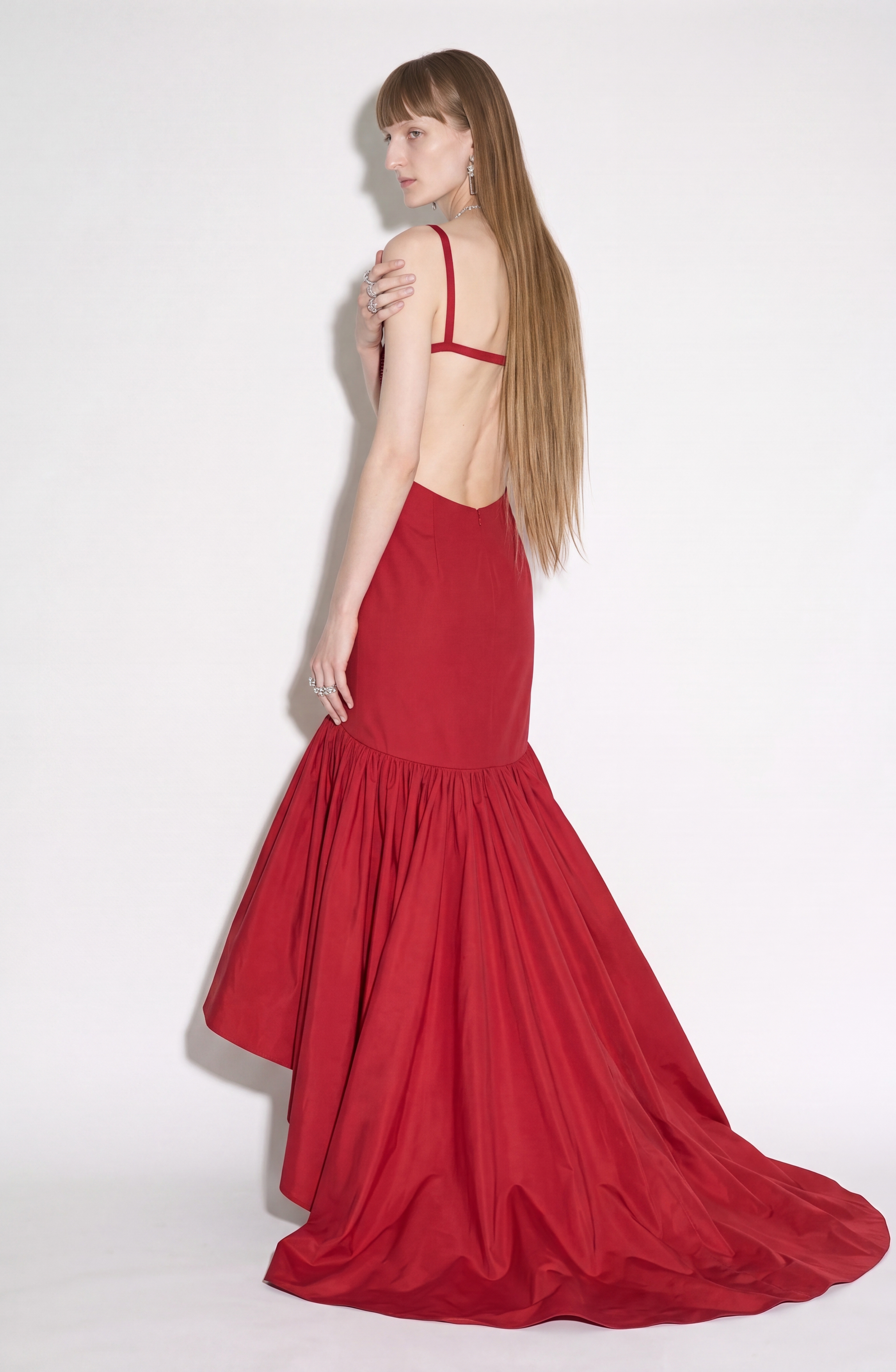 A woman with long, straight hair wears the LOULOU Crystal-Embellished Bustier Gown, featuring a stunning ruby red hue, backless design, and dramatic ruffled train. She stands against a plain white background, looking over her shoulder.