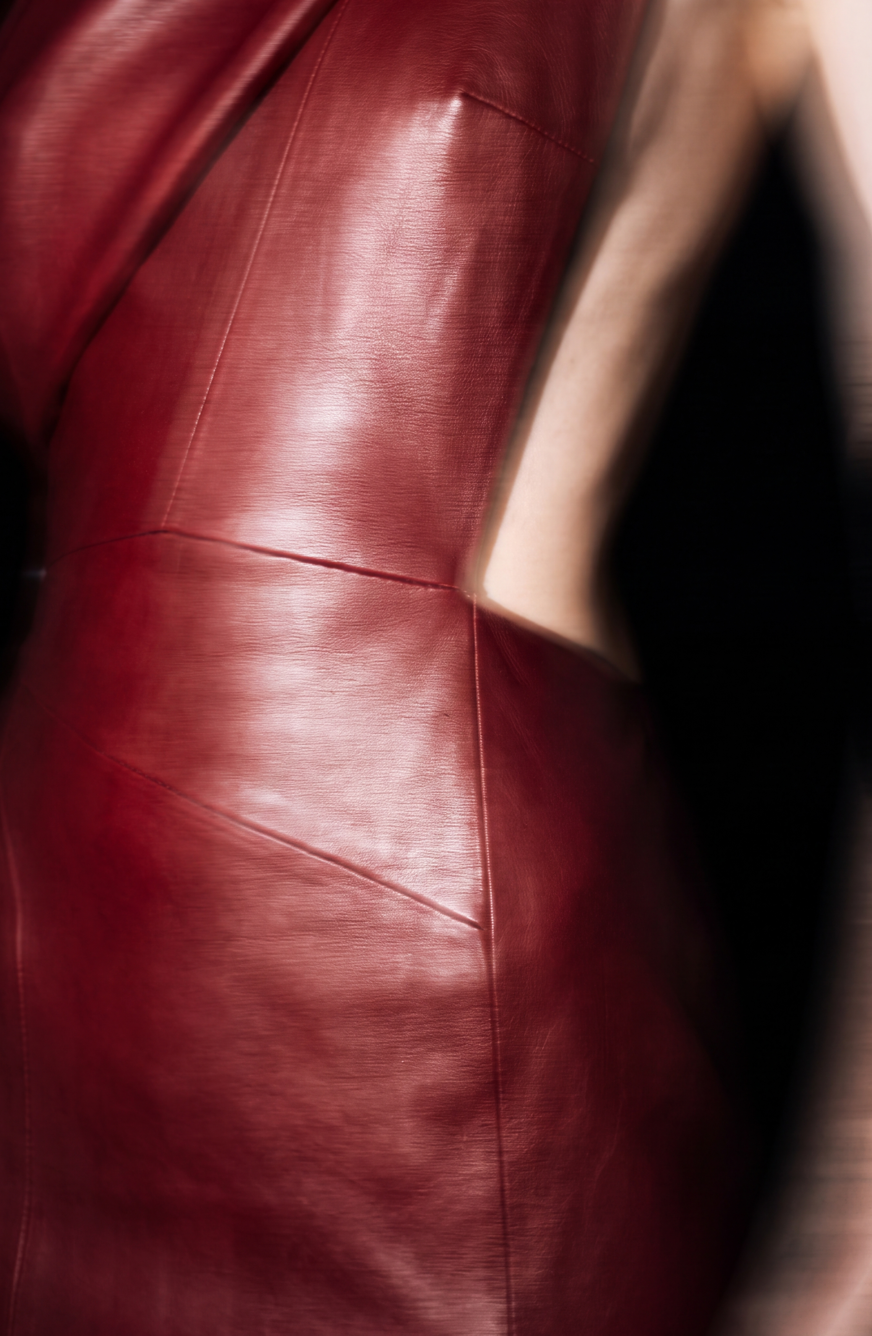 A close-up of the LOULOU One-Shoulder Leather Maxi Dress in glossy dark red oxblood, featuring cut-out sides that reveal part of the wearer’s bare back and arm, set against a dark background.