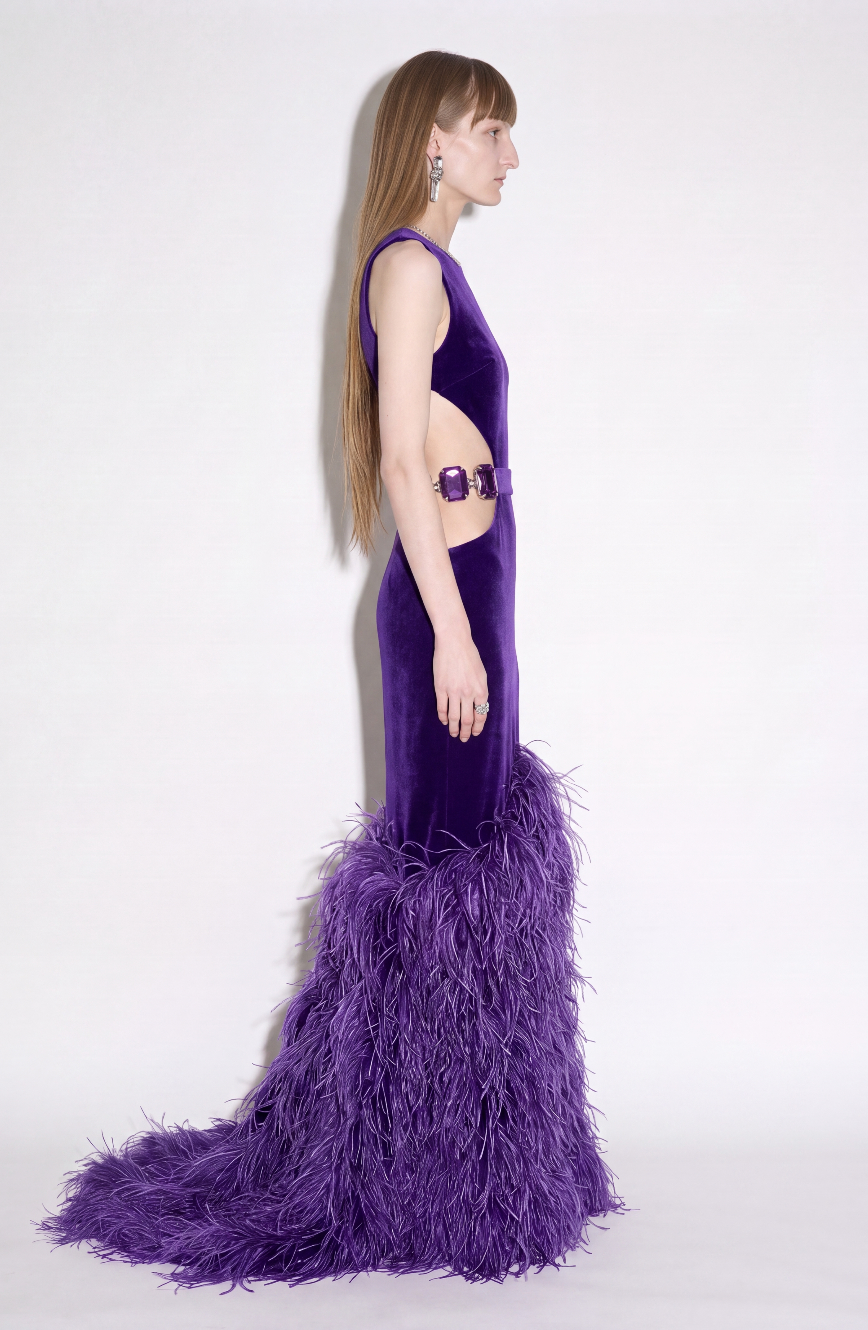 A woman models the LOULOU Velvet Cutout Feather Gown, a sleeveless, floor-length evening dress featuring a feather-trimmed skirt and side cutout, as she stands in profile against a plain white background.