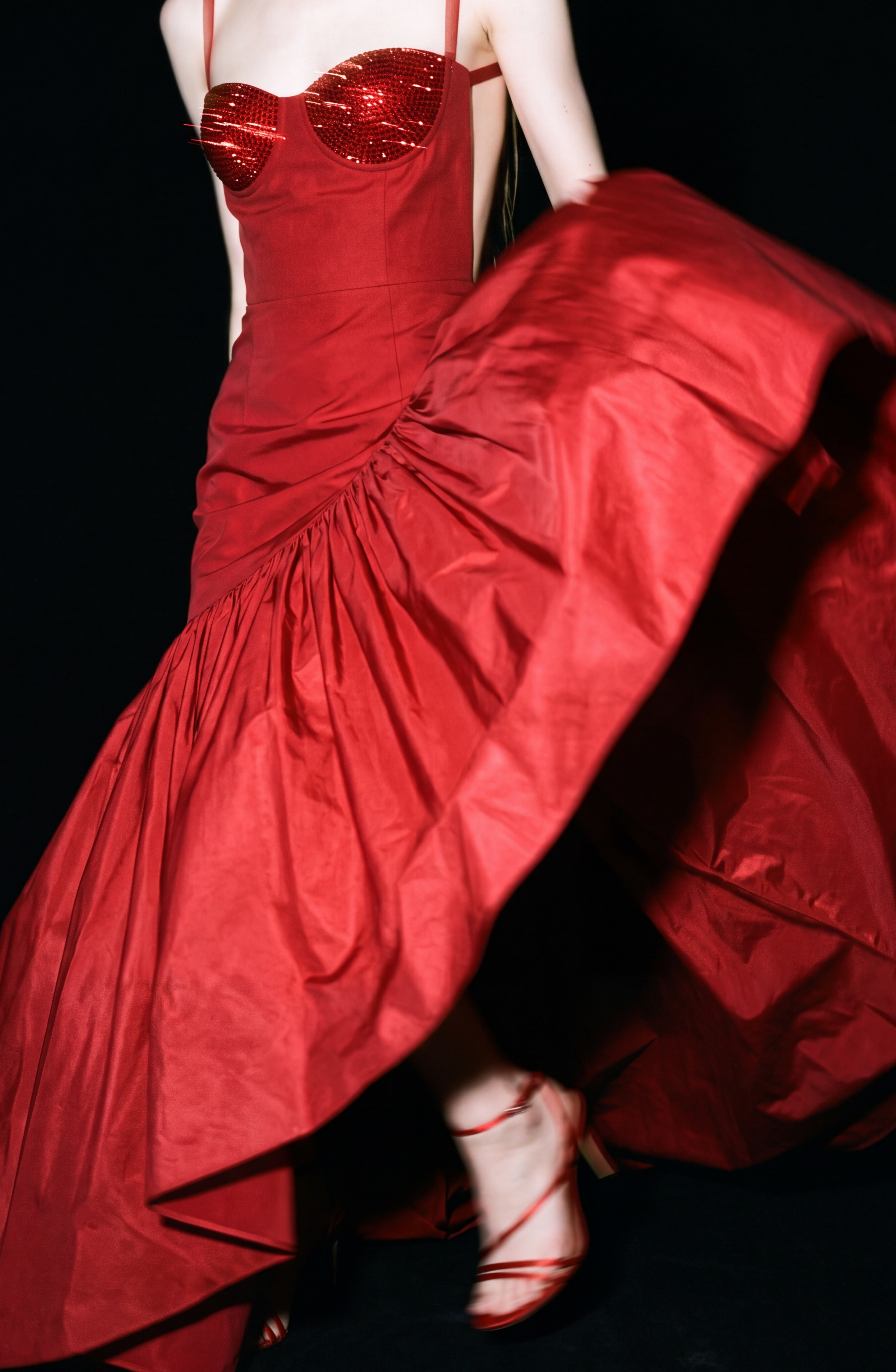 A person twirls in the LOULOU Crystal-Embellished Bustier Gown—a flowing, strapless ruby red dress with a sparkling crystal-adorned bodice—causing the skirt to flare dramatically against a black background that highlights both the gown and the movement.