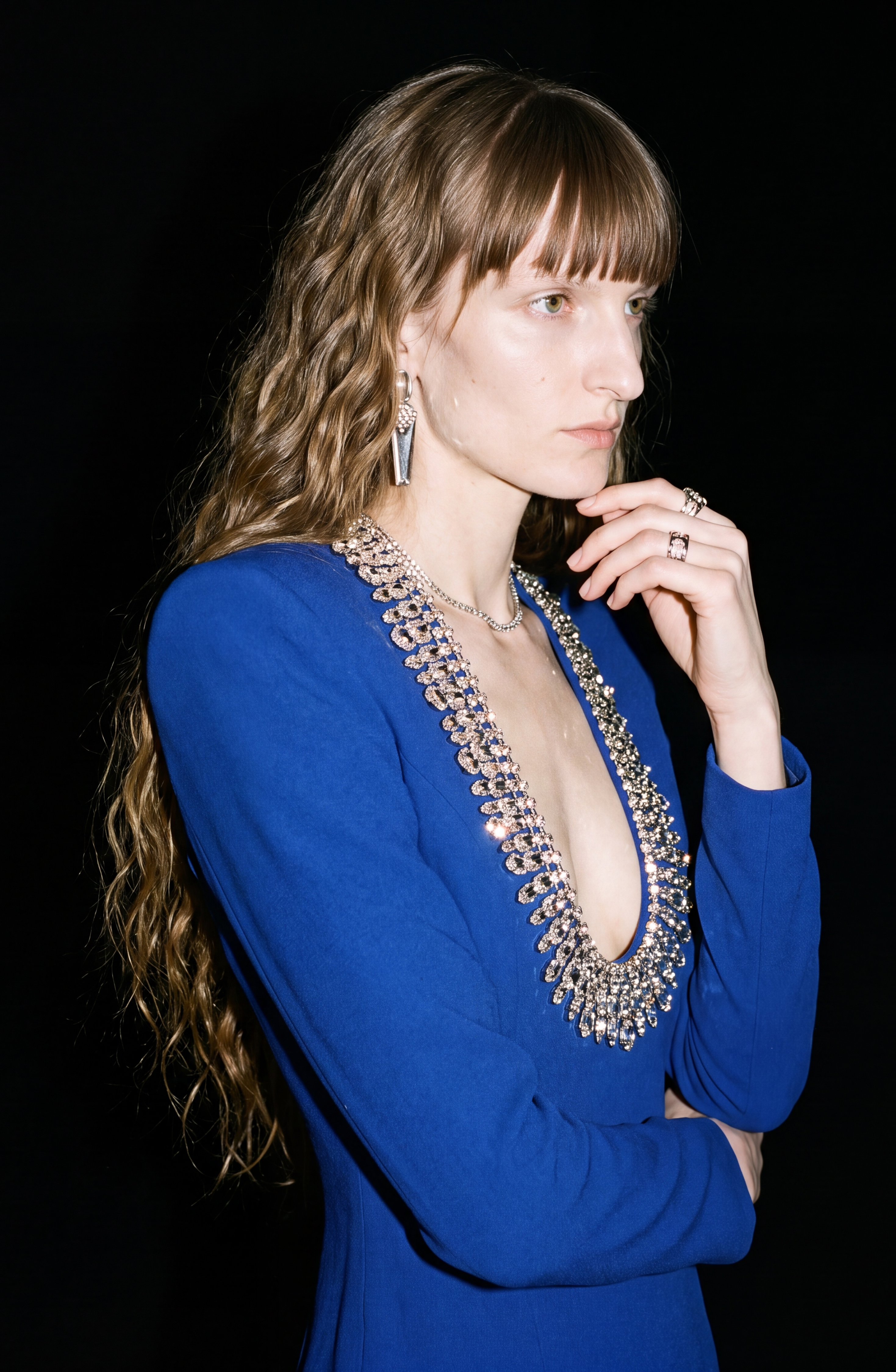 A person with long wavy hair poses thoughtfully against a black background, wearing the Crystal U-Neck Midi Dress by LOULOU—featuring crystal embellishments and a jeweled neckline in deep blue—with one hand near their chin and the other arm crossed.
