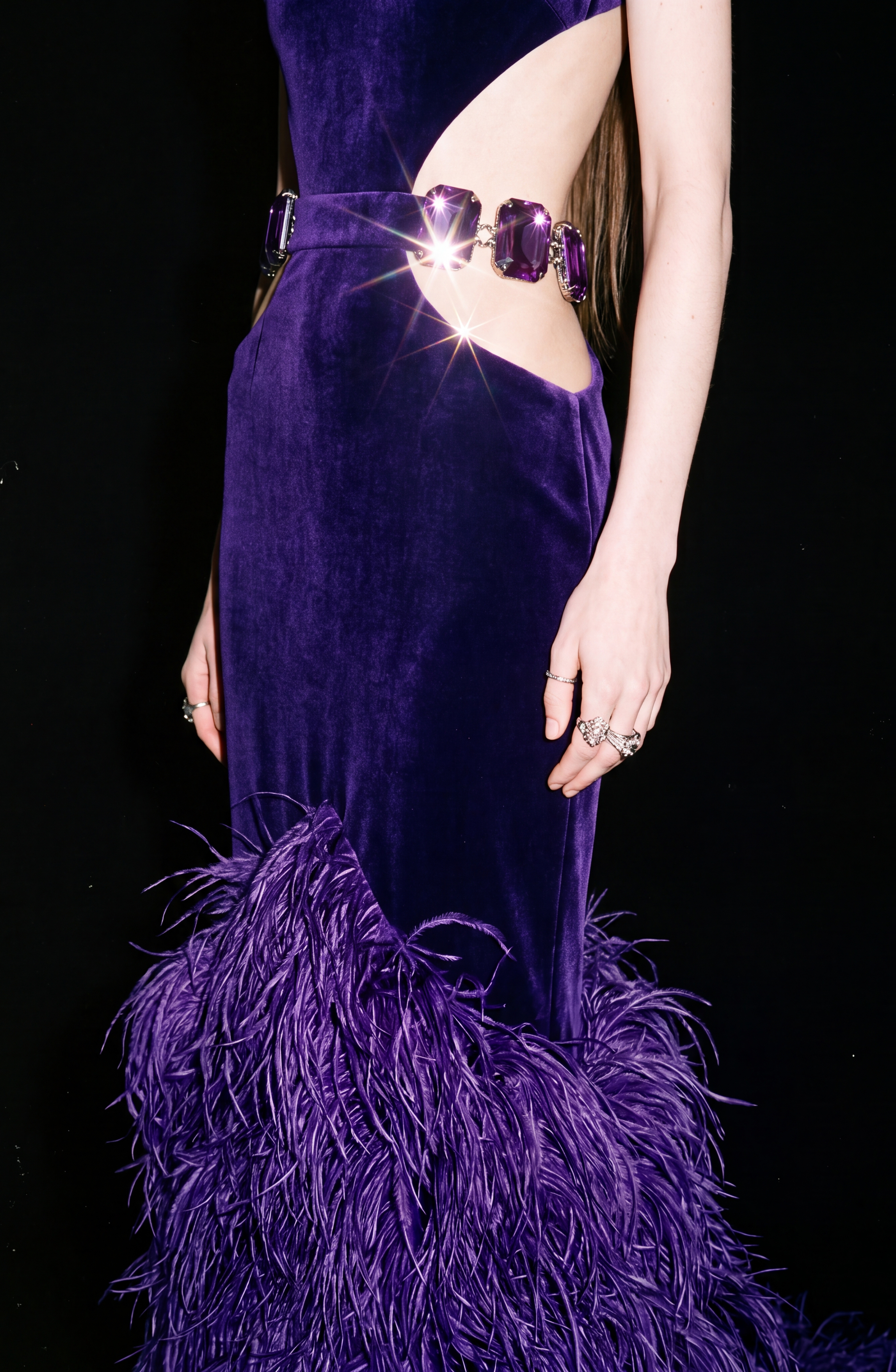 A close-up view of a person wearing the LOULOU Velvet Cutout Feather Gown, a vibrant purple evening dress featuring bold side cutouts connected by large crystal buckles. The gown is finished with a dramatic feather-trimmed skirt, and the model’s hands are adorned with several ornate rings.