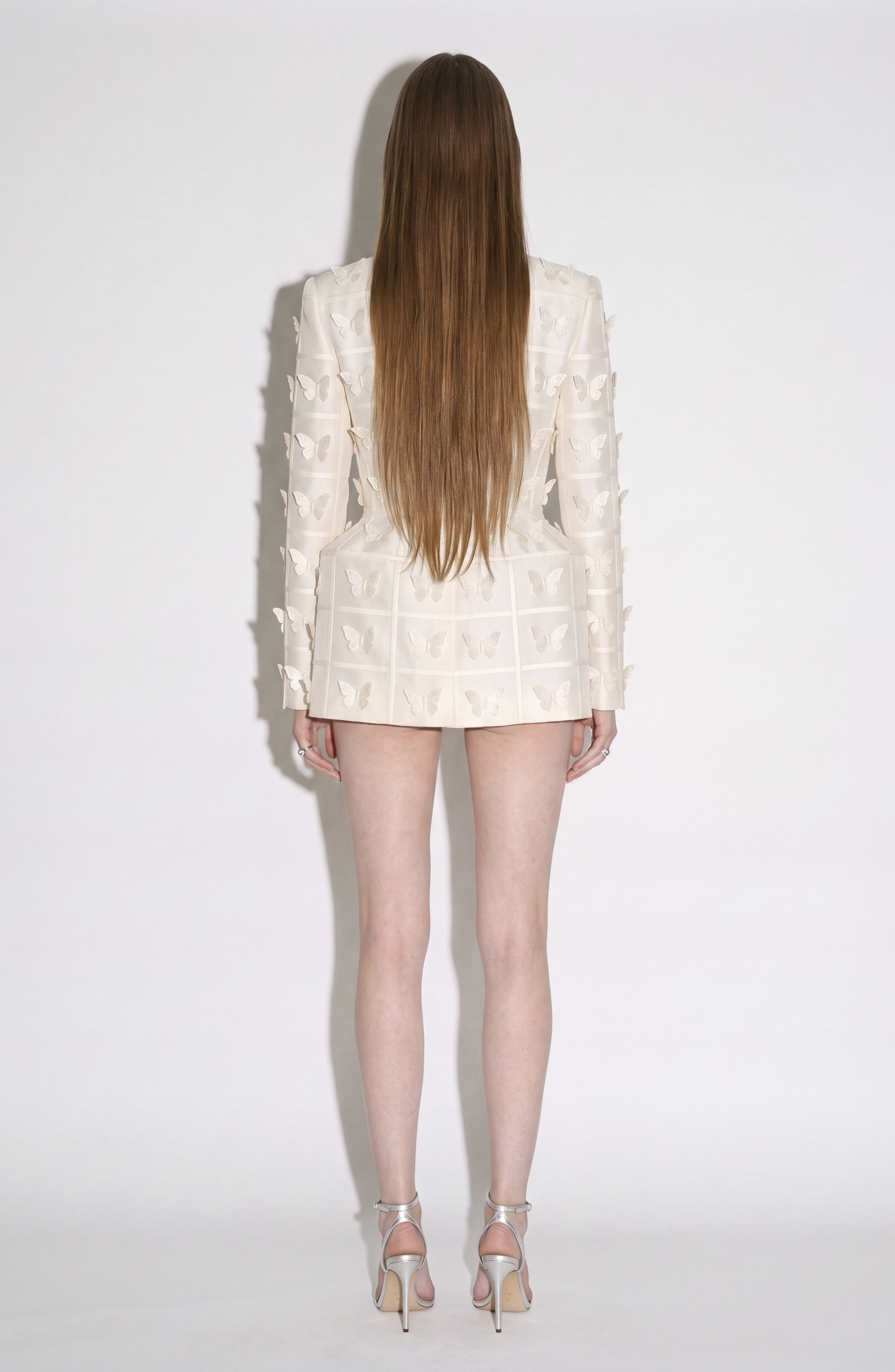 A person with long straight hair stands facing away, wearing the LOULOU Embellished Silk Basque Blazer Dress, featuring a white textured finish and tailored silhouette paired with high heels, against a plain light background.