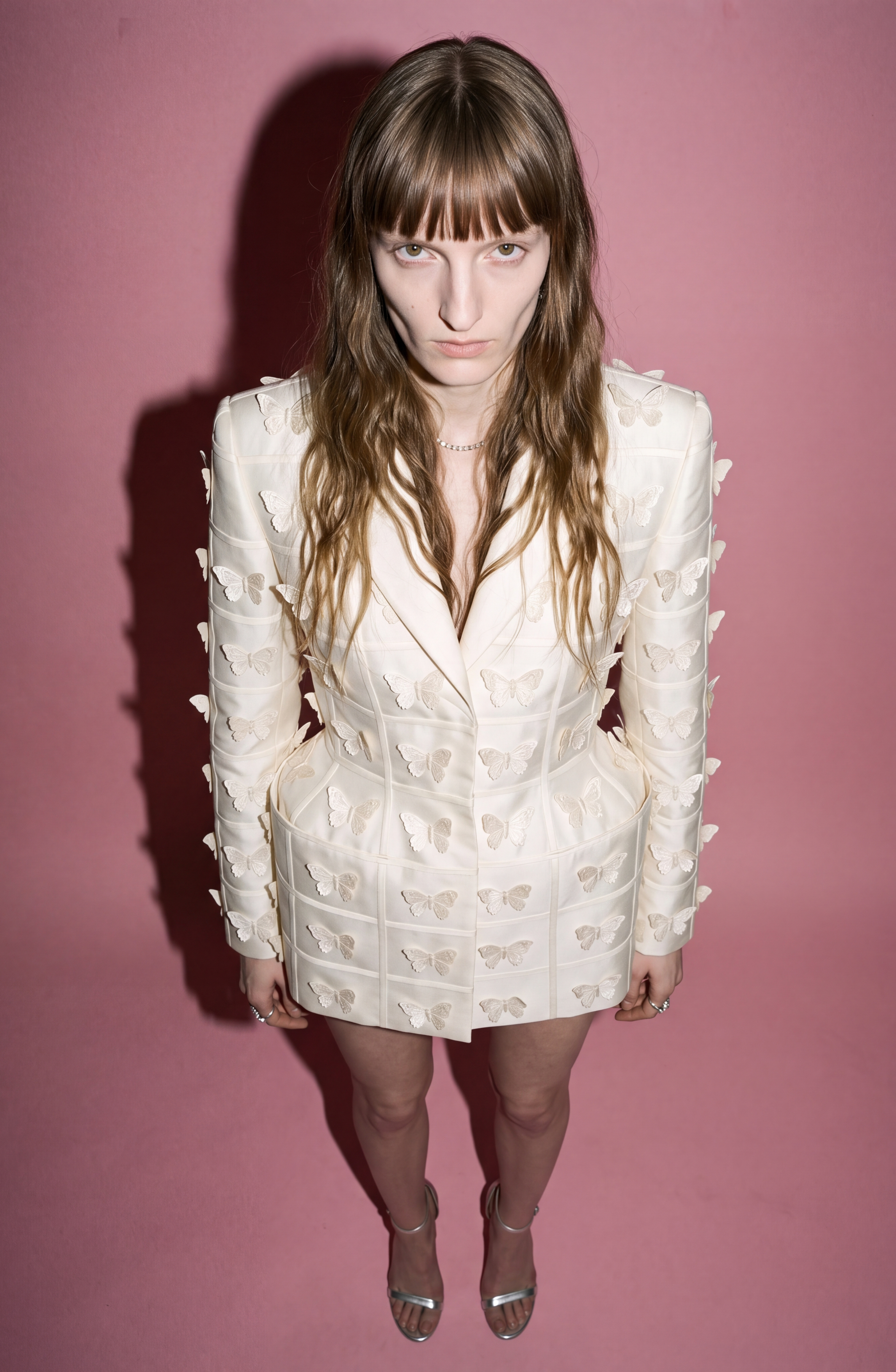 A person with long hair stands against a pink background, wearing the LOULOU Embellished Silk Basque Blazer Dress in ivory, featuring delicate butterfly appliqués and styled with silver heels. With a serious expression, they face the camera as the dress’s tailored silhouette enhances its sophisticated appeal.