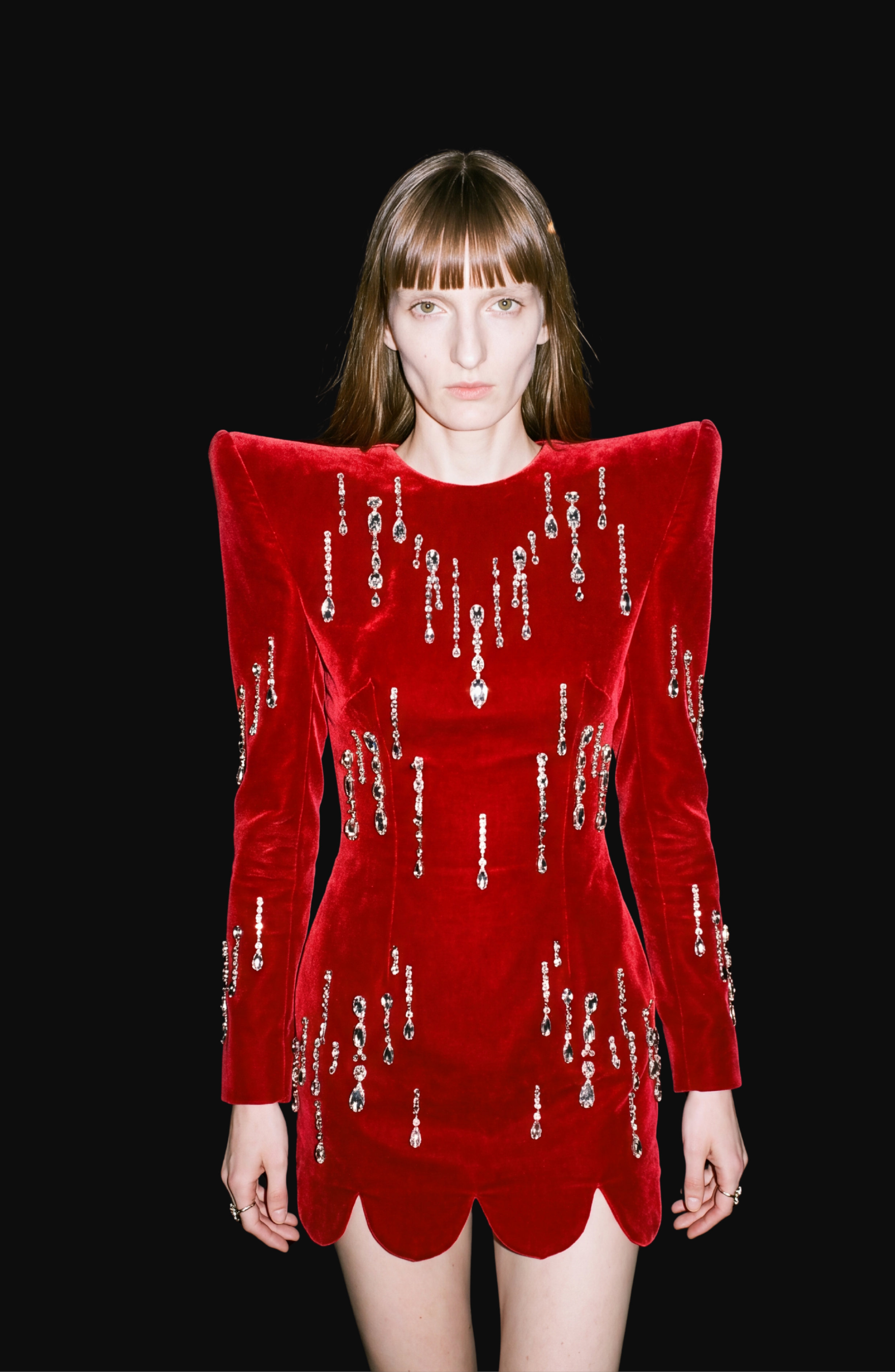 A person with straight brown hair models the LOULOU Scarlet Velvet Mini Dress, featuring sculptural shoulders and glittering glass crystal embellishments, posed against a plain black background.