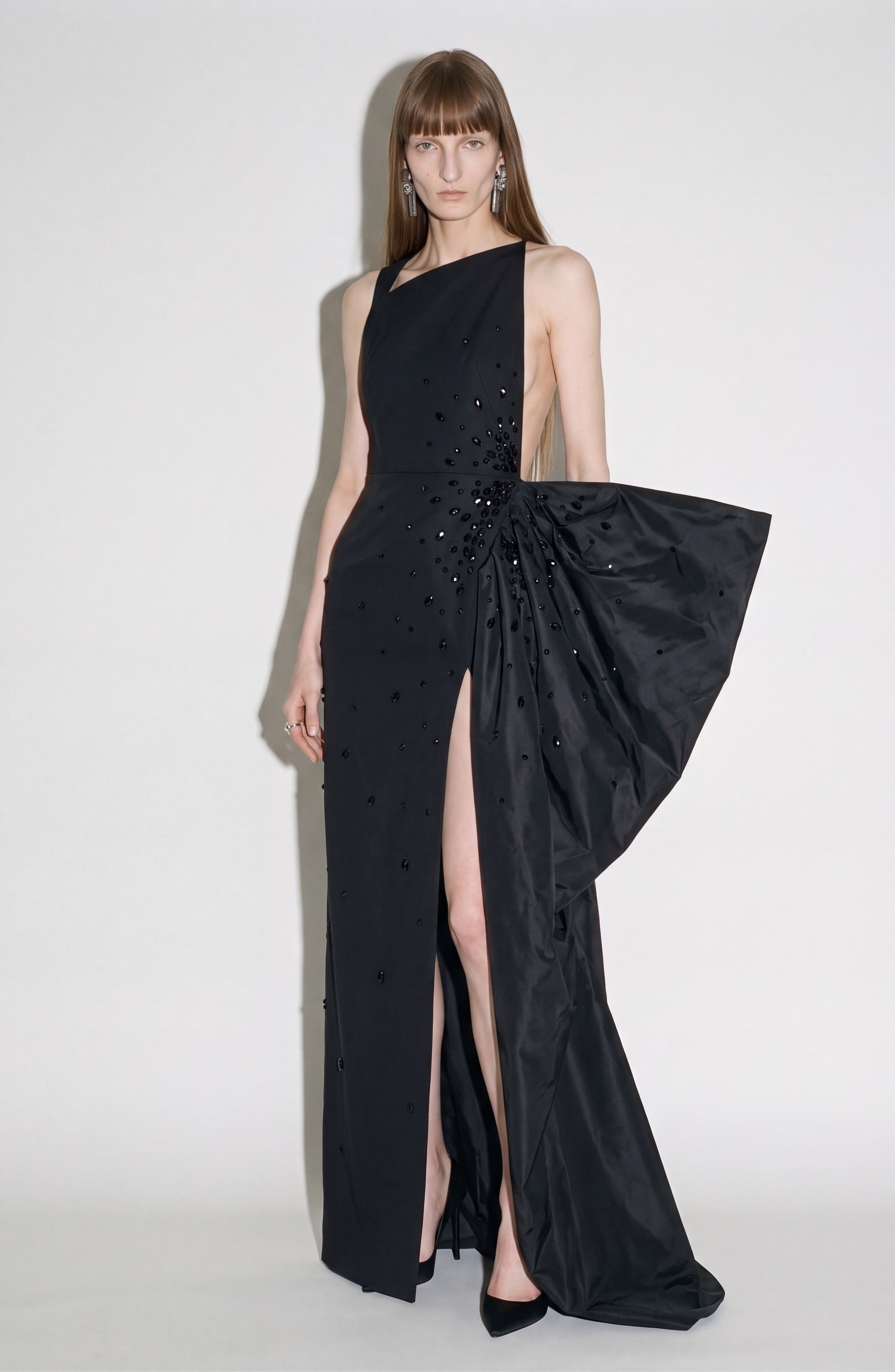 A woman showcases the LOULOU Asymmetric Crystal Bow Gown—a sleek, jet-black dress with an asymmetric neckline, bold side slit, sparkling crystal embellishments, and a dramatic oversized bow at the side—paired with black heels against a plain background.