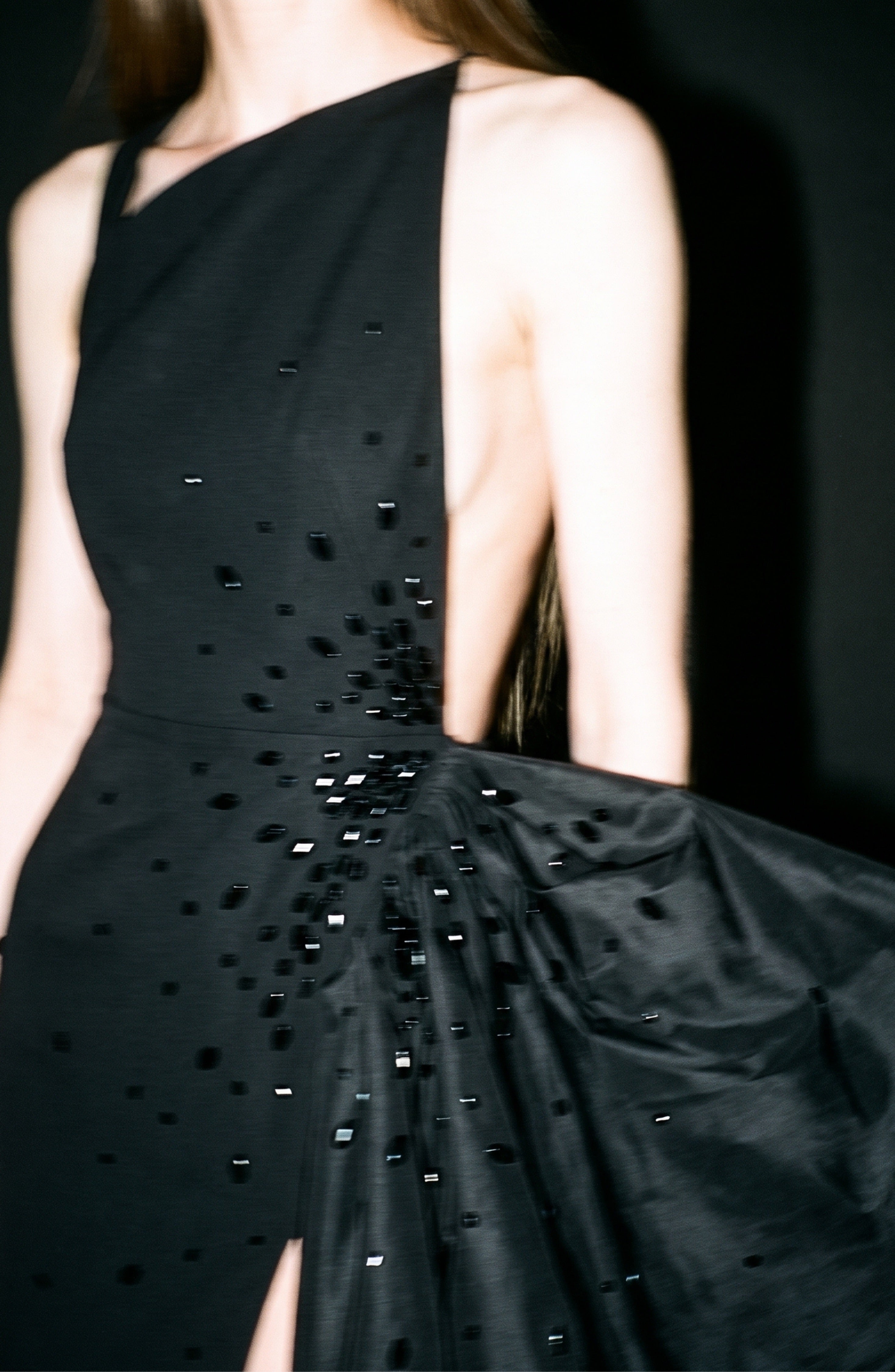 A person stands against a dark background, wearing the LOULOU Asymmetric Crystal Bow Gown—a jet black dress adorned with shiny crystal embellishments, a dramatic side drape, and an asymmetric neckline. The image appears slightly blurred.