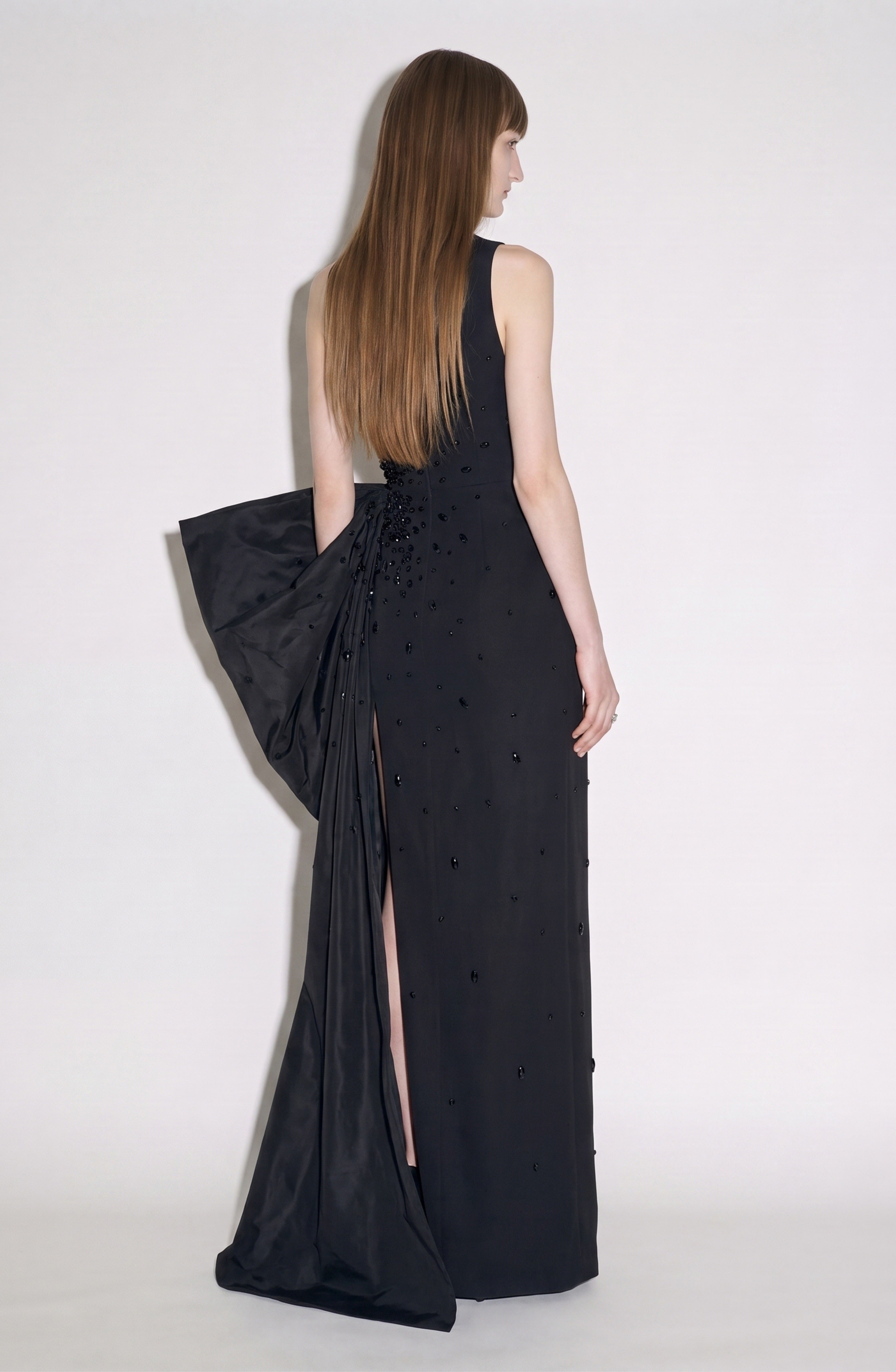 A woman with long straight hair models the LOULOU Asymmetrical Embellished Gown—a sleeveless black design featuring a striking bow and back slit, its sleek silhouette and elegant asymmetrical neckline showcased against a plain white backdrop.