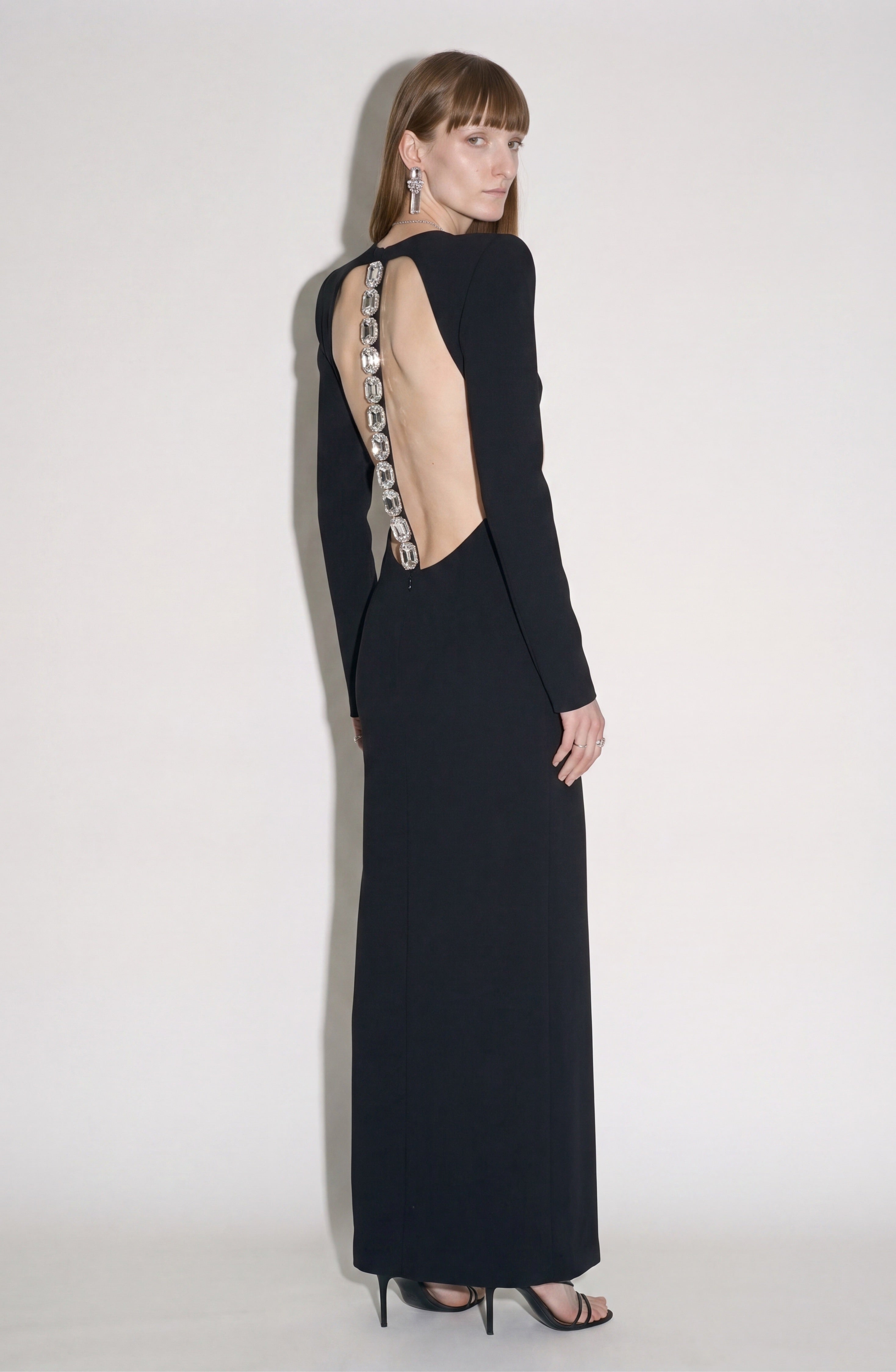 A woman wearing the LOULOU Back Crepe Maxi Dress—featuring a long black silhouette with metallic detailing along the spine—stands sideways against a plain light background, glancing over her shoulder. She completes the elegant look with earrings and high heels, while an open back adds an alluring touch.