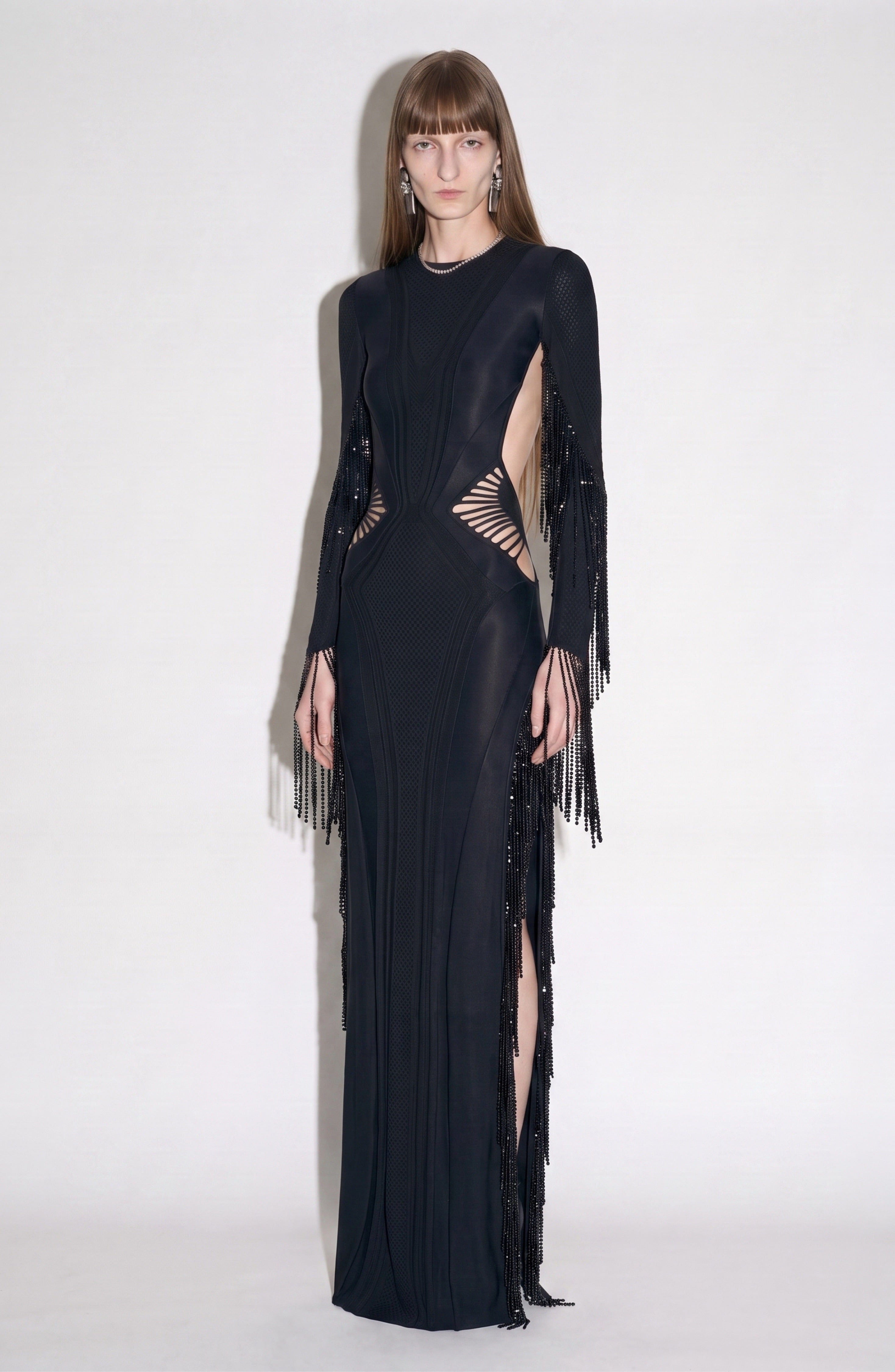 A person wearing the LOULOU Beaded Fringe Cut-Out Gown, featuring a jet black hue, geometric paneling, side cutouts, and intricate beaded fringe detailing, stands against a plain white background facing forward.