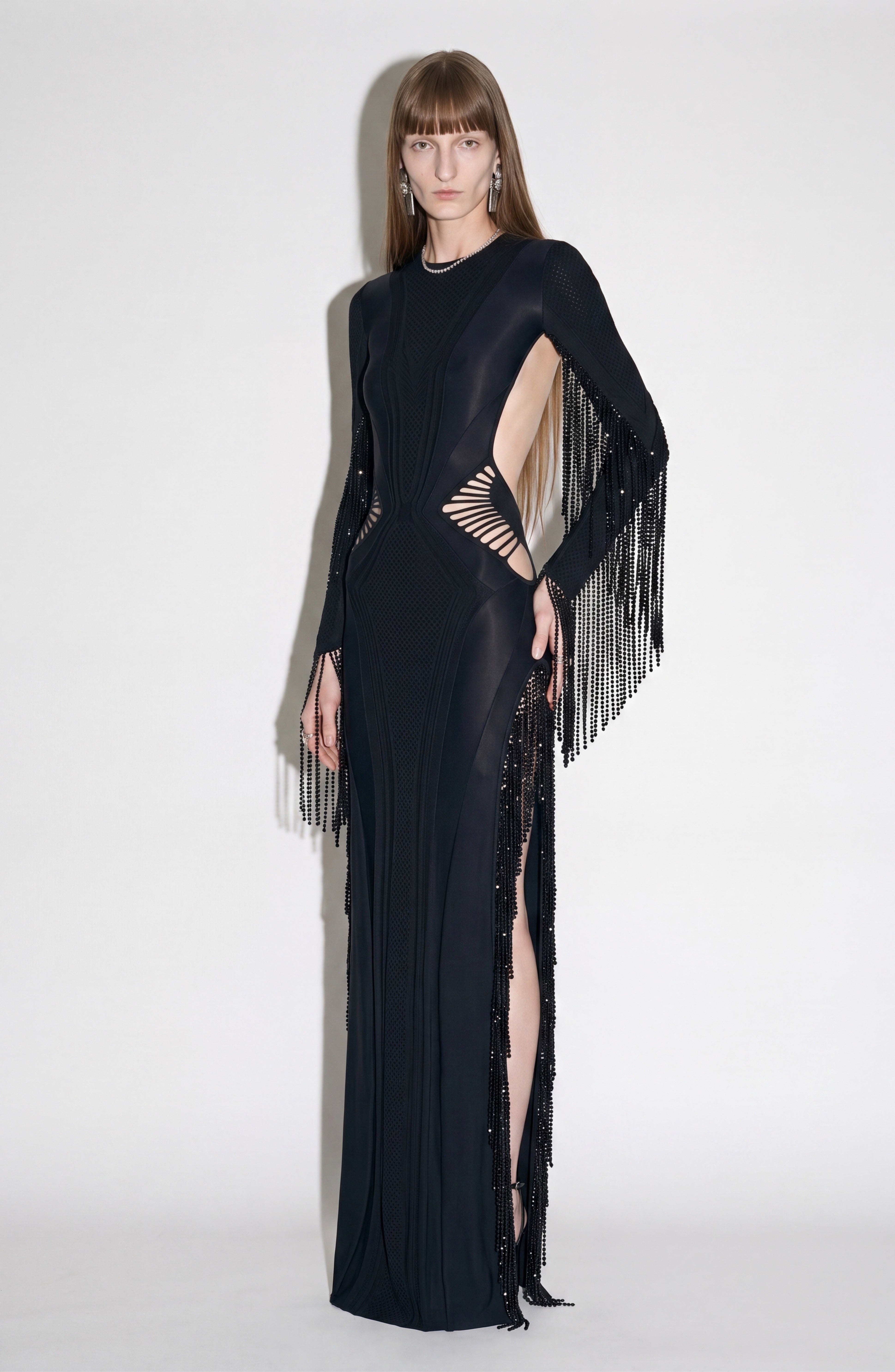 A person wearing the LOULOU Beaded Fringe Cutout Maxi Dress, a long black gown with geometric cutouts, beaded fringe detailing on the sleeves, and a high side slit, stands against a plain white background.