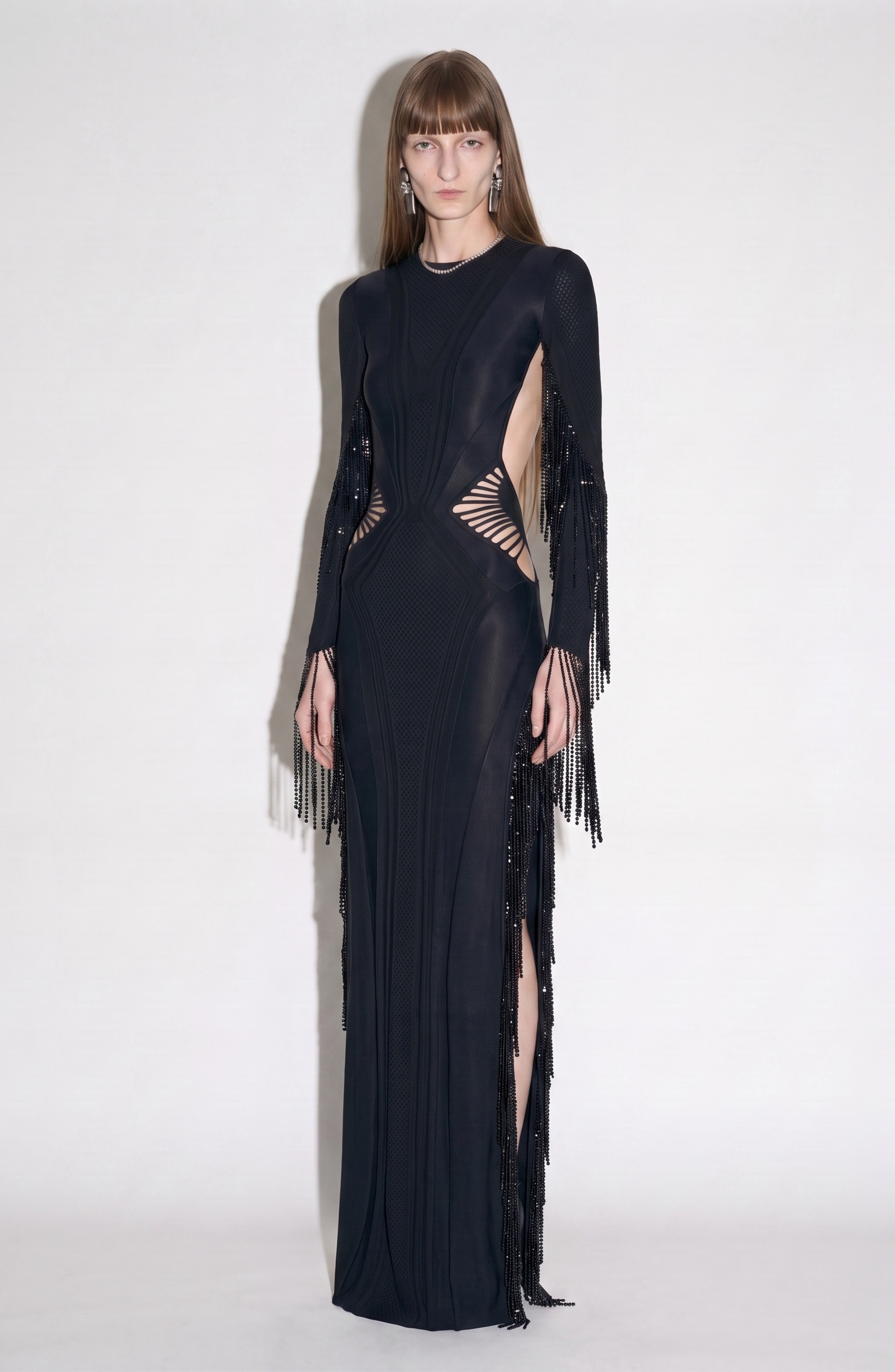 A person stands against a plain background wearing the LOULOU Beaded Fringe Cutout Maxi Dress, featuring geometric cutouts, long sleeves, and beaded fringe details, looking directly at the camera.