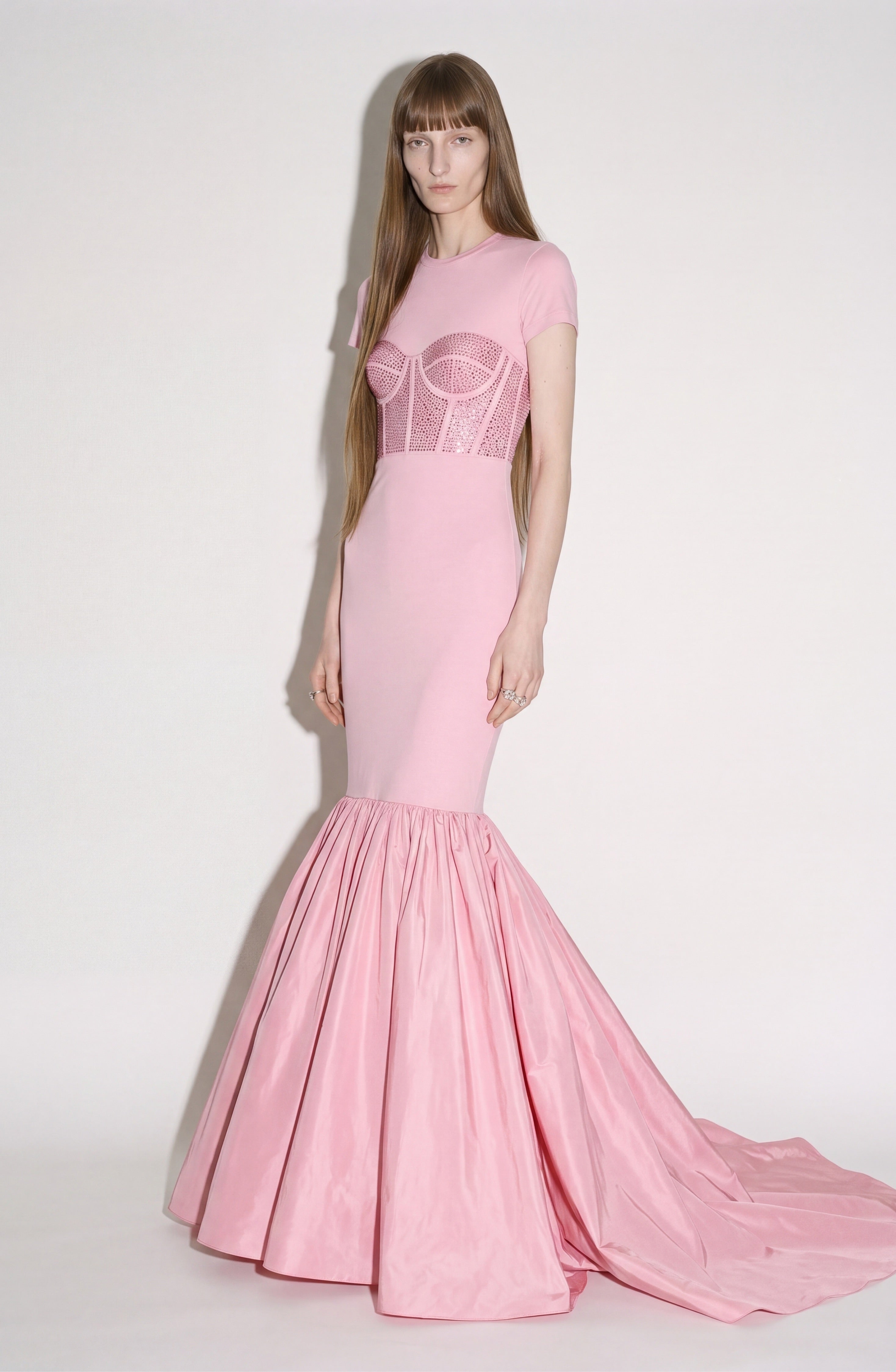 A person stands in the LOULOU Corset Mermaid Gown—a fitted, pink design featuring a crystal-embellished corset and a dramatic flared taffeta skirt—set against a plain light background.