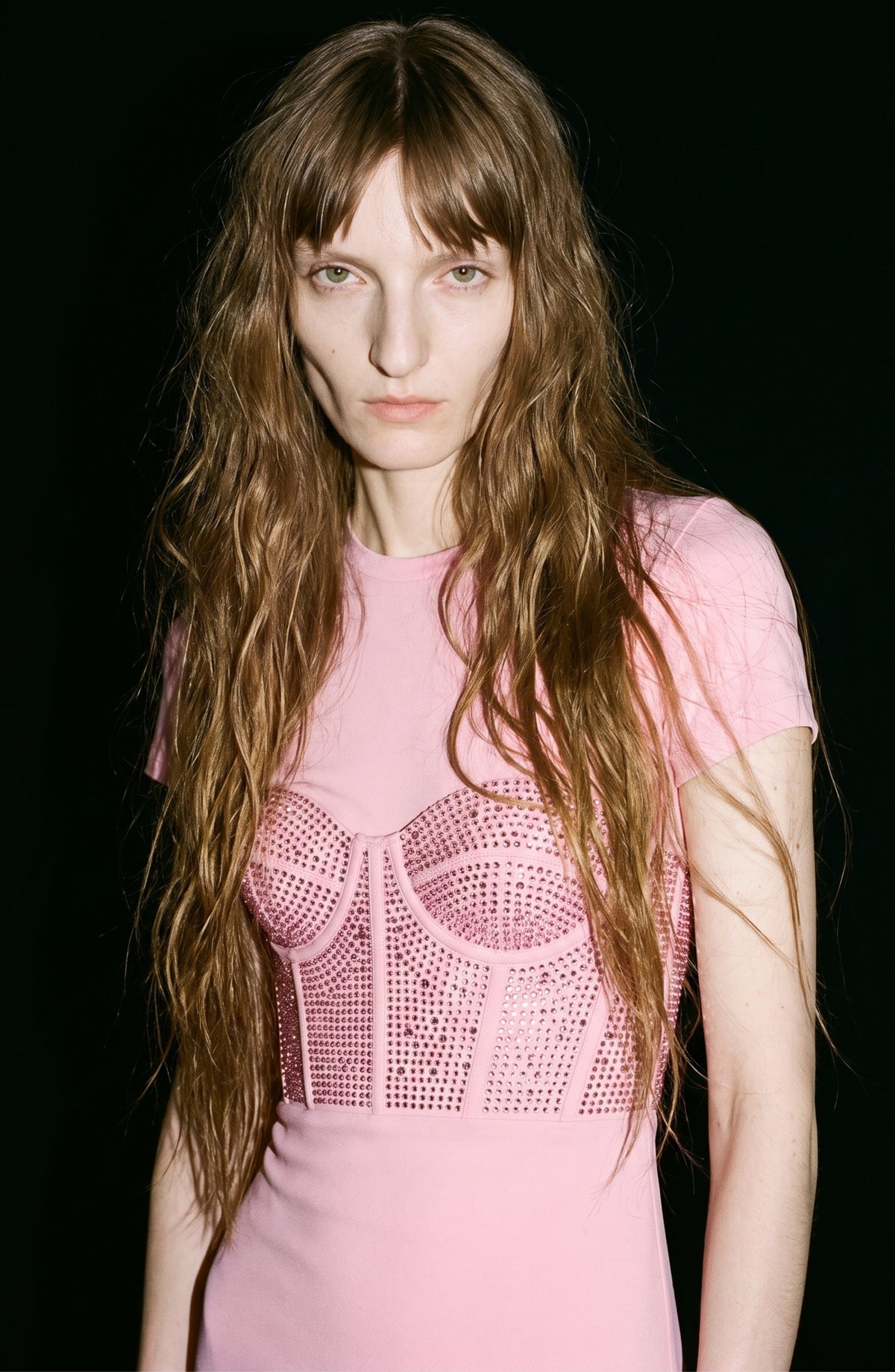 A person with long wavy hair wears the LOULOU Corset Mermaid Gown in light pink silk taffeta, featuring decorative stud details on the bodice, standing against a black background.