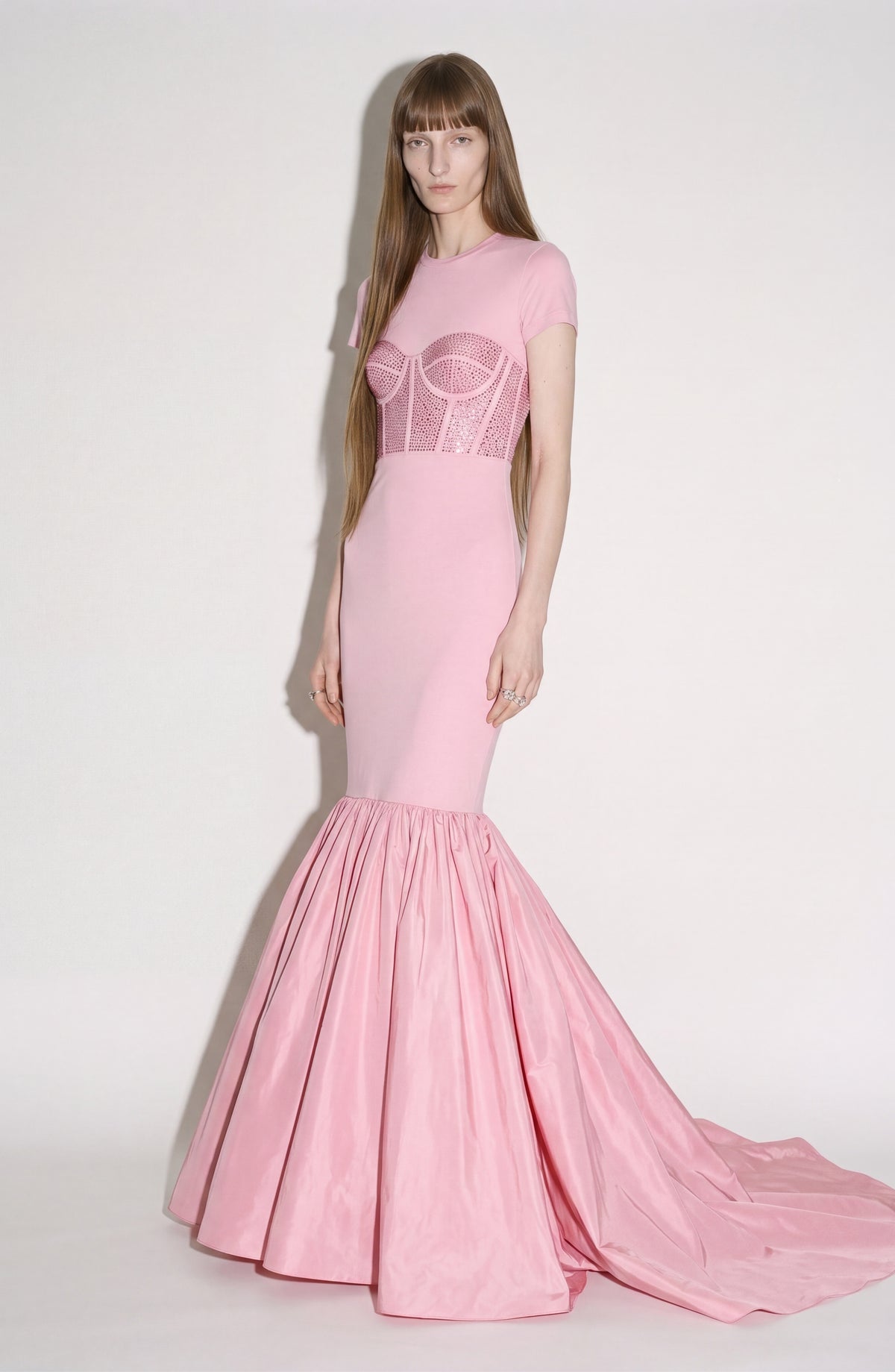 A model stands against a plain white background wearing the LOULOU Corset Mermaid Gown—a fitted, light pink dress crafted from luxurious silk taffeta with a structured bodice and a flared, floor-length skirt.