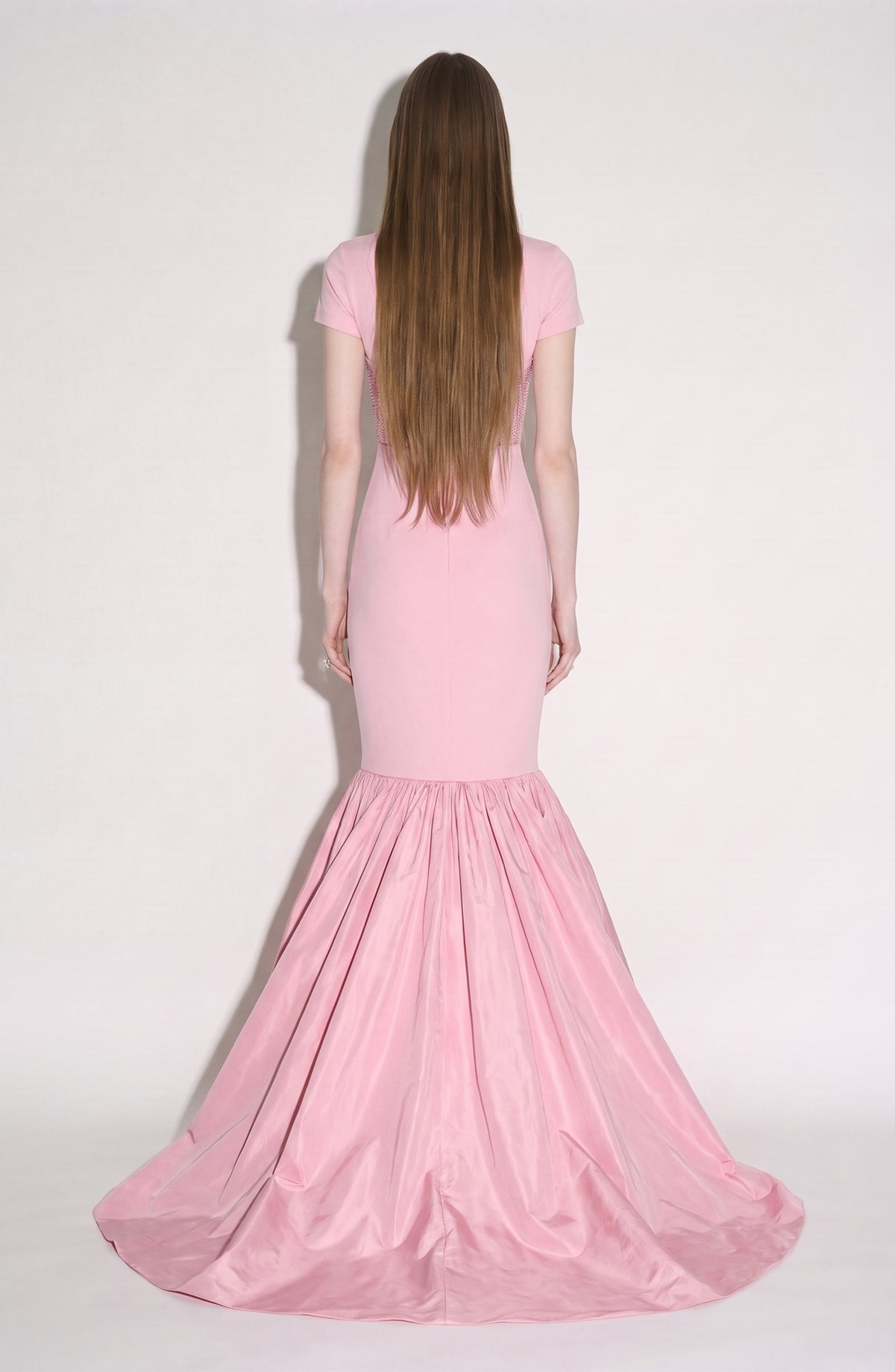 A woman with long brown hair stands facing away, wearing the LOULOU Corset Mermaid Gown—a fitted pink mermaid silhouette crafted from luxurious silk taffeta, its flared skirt elegantly pooling on the floor.