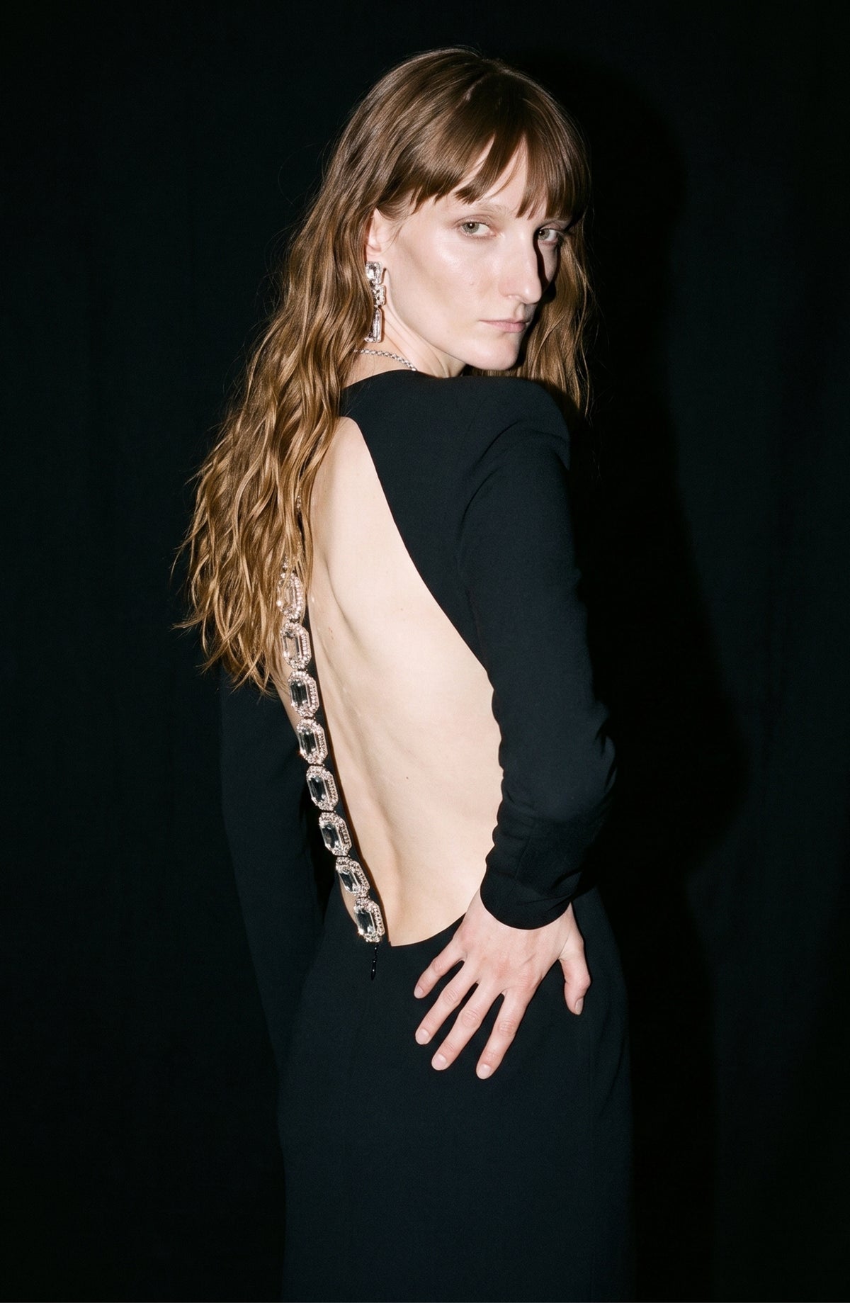 A person with long wavy hair poses against a dark background, wearing the LOULOU Crepe Maxi Dress—a black maxi dress with a dramatic open back and jewel-like embellishments accenting the spine.