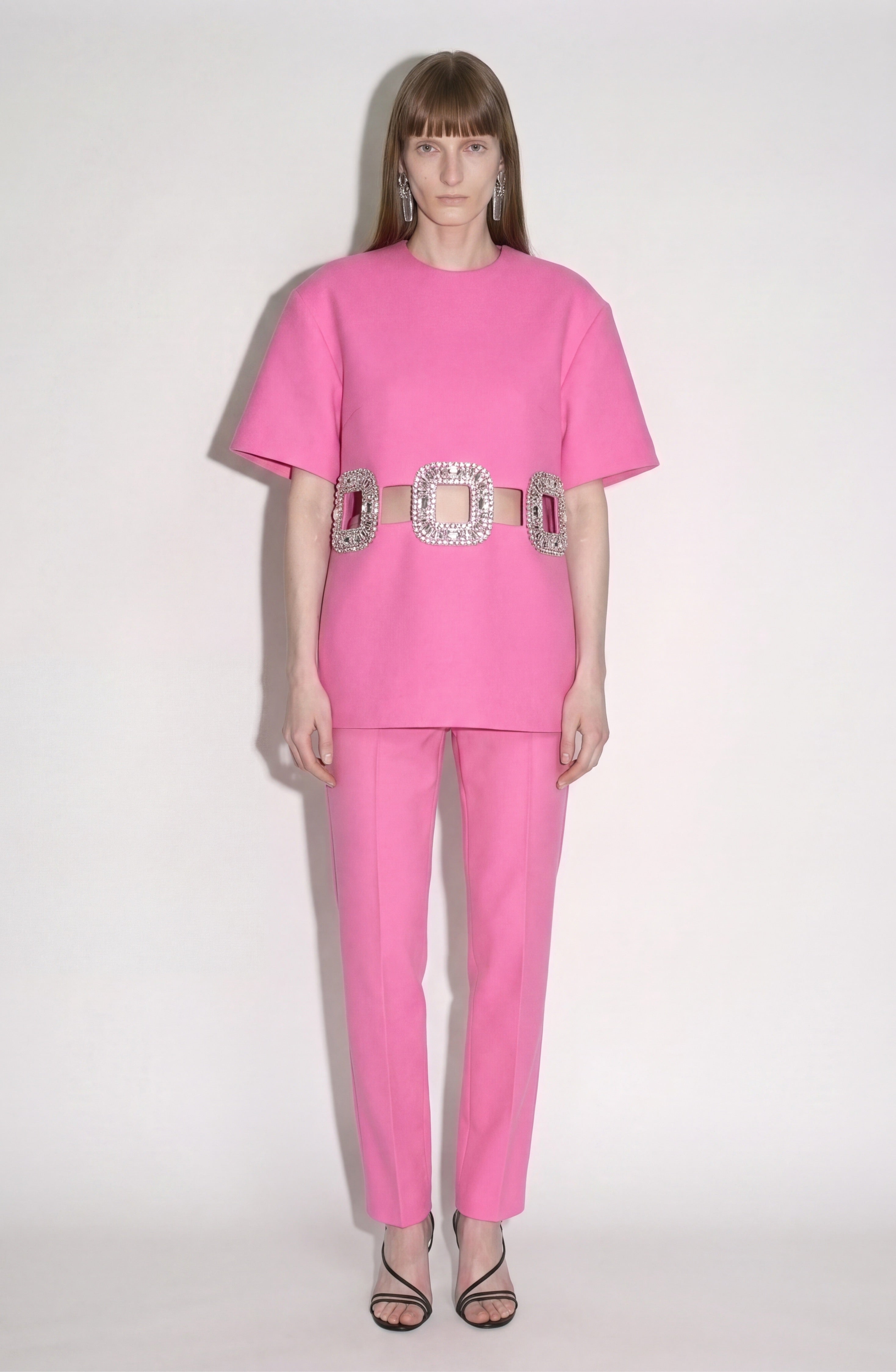 A person stands against a plain background wearing a bubblegum pink outfit featuring LOULOU’s Cropped Tailored Trousers, paired with a large, sparkly square-buckle belt and black strappy heels.