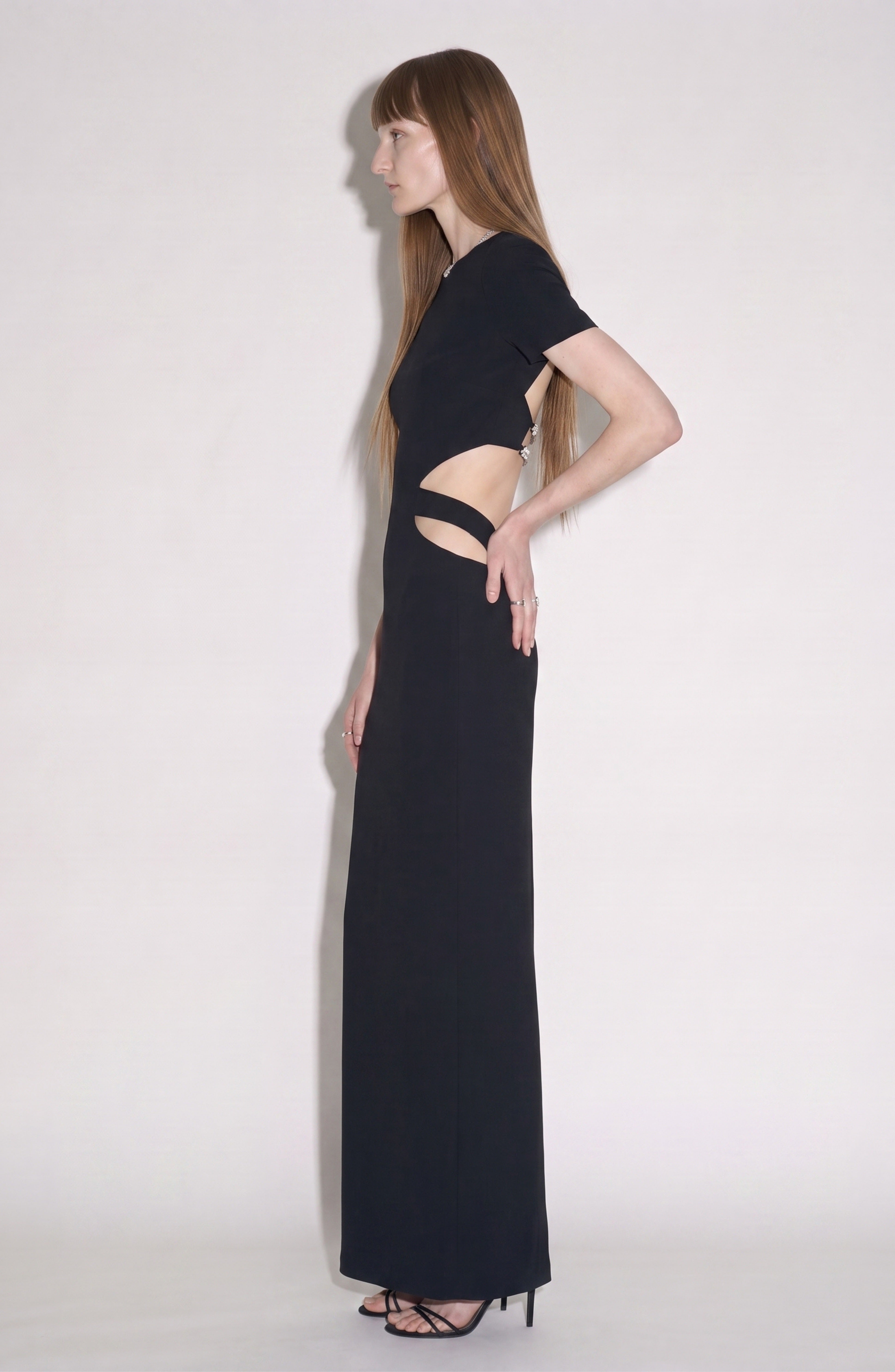 A woman stands in profile against a plain white background, wearing the LOULOU Crystal-Embellished Cutout Maxi Dress with sleek black high heels.