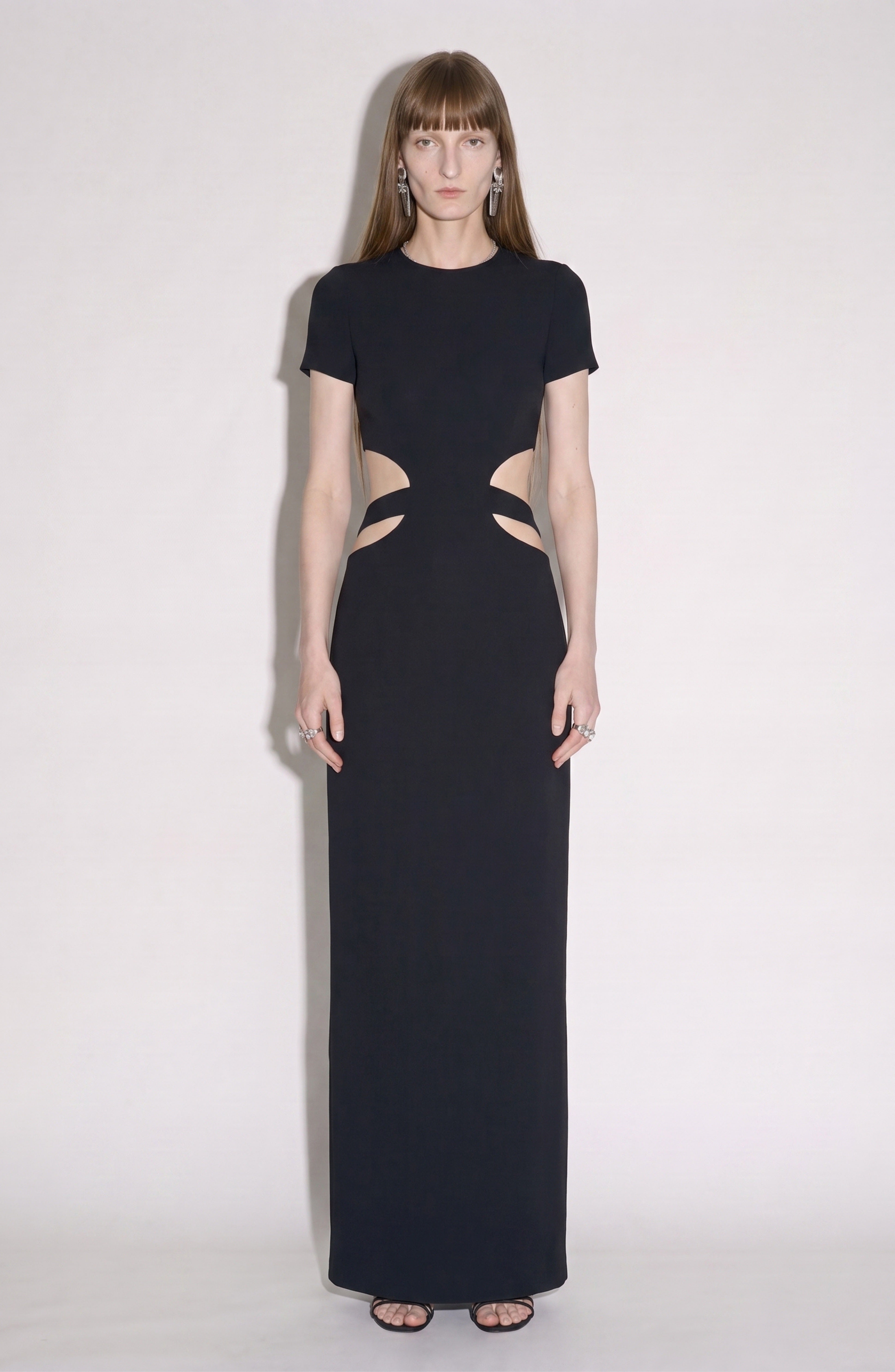 A woman stands against a plain background wearing the LOULOU Crystal-Embellished Cutout Maxi Dress, featuring short sleeves and stylish side cutouts at the waist, paired with black open-toe heels.