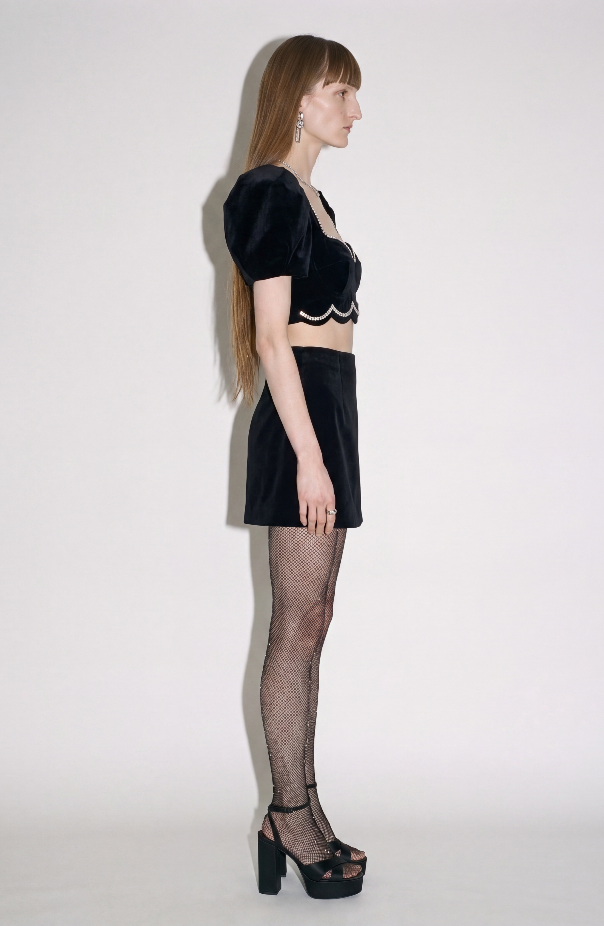 A person with long hair stands in profile against a plain white background, wearing the LOULOU Crystal-Trim Velvet Crop Top—a black velvet bustier with puff sleeves—paired with a black mini skirt, fishnet stockings, black platform heels, and long earrings.