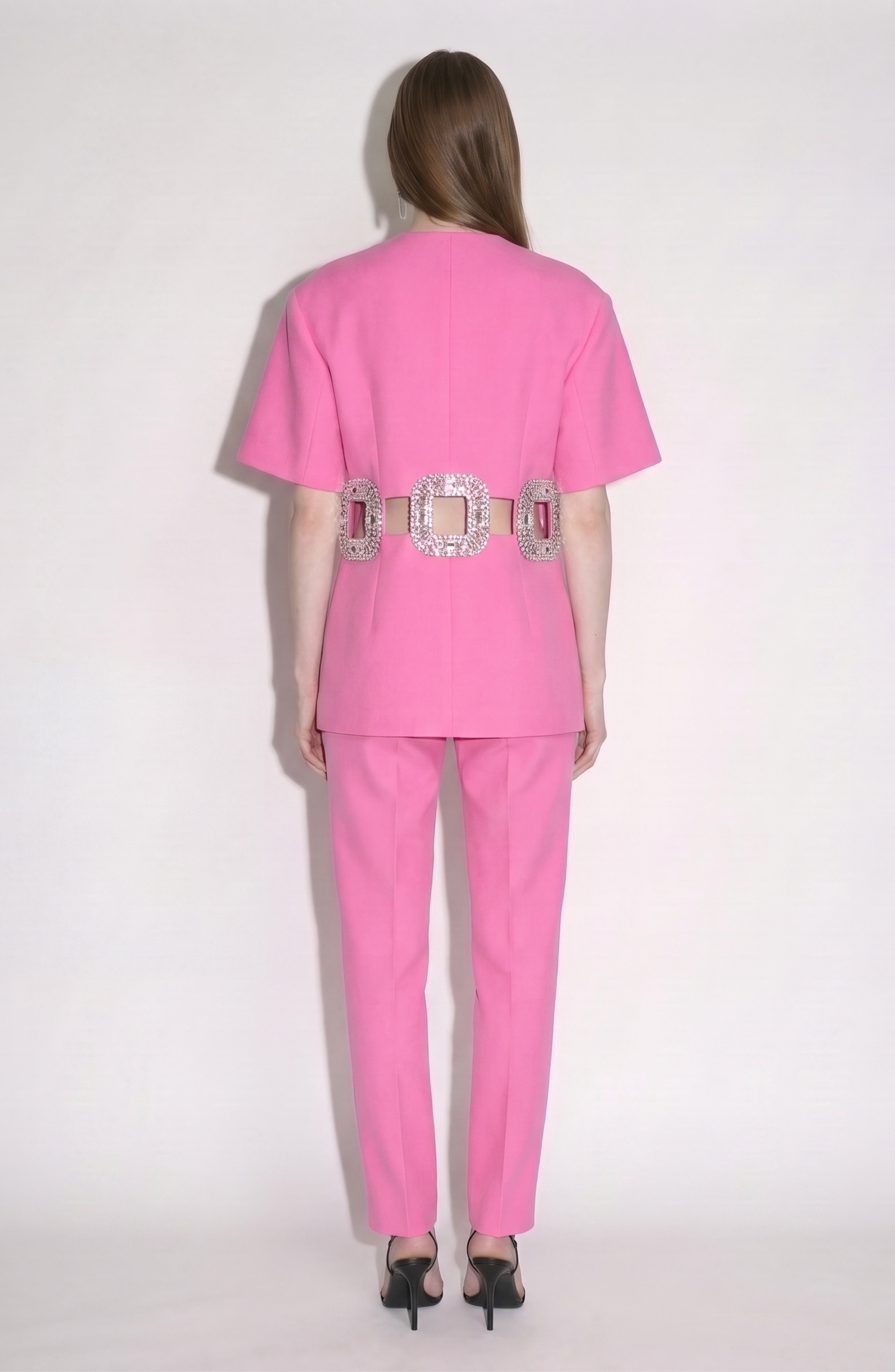 A woman stands facing away, wearing LOULOU's Crystal Buckle Cut-Out Top—a bright pink, short-sleeved blazer with a wide, crystal-embellished buckle belt at the waist—paired with matching pants and black high-heeled sandals.