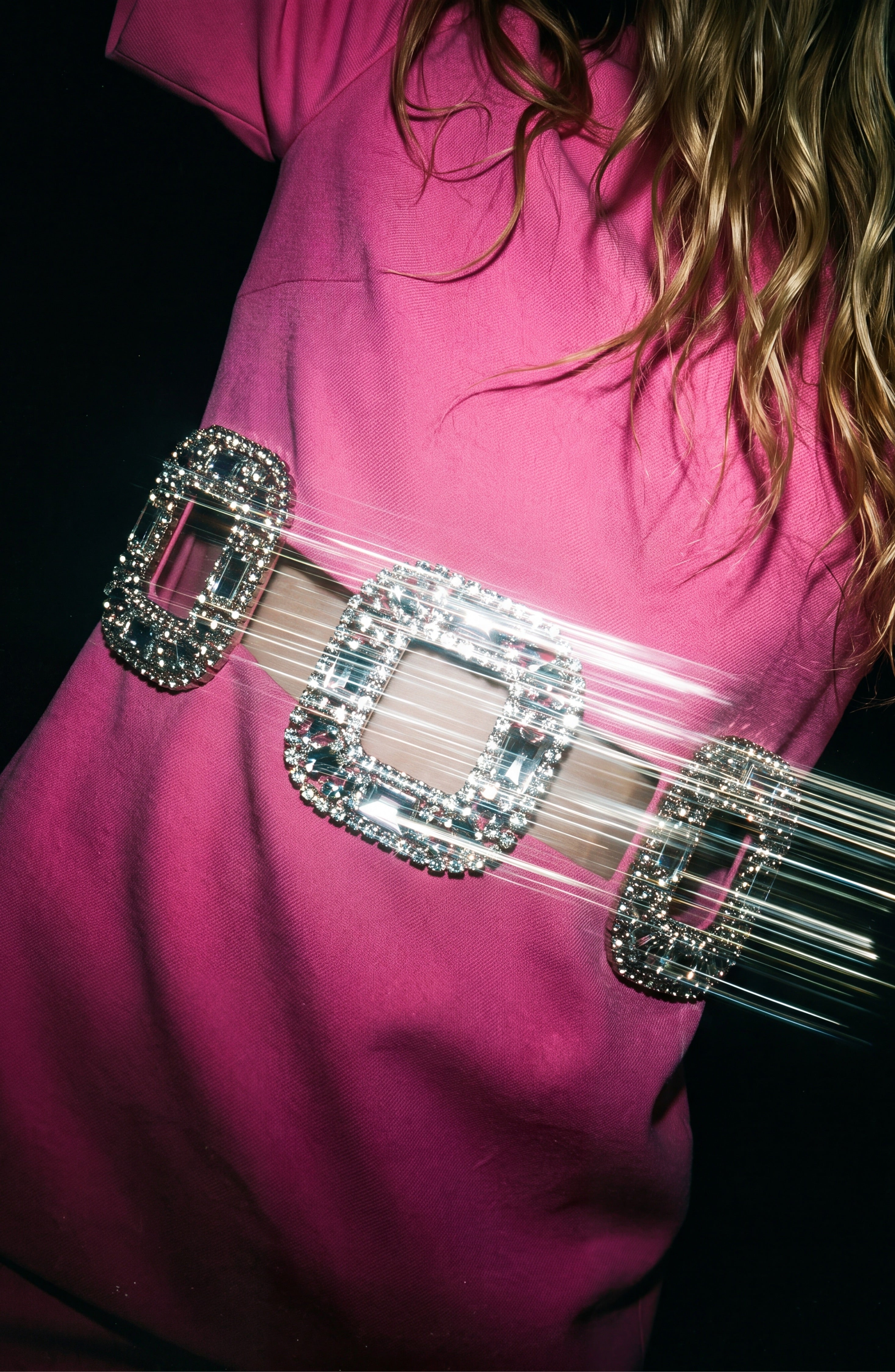 A person in a bright pink Crystal Buckle Cut-Out Top by LOULOU, with wavy hair, wears the top featuring large, shiny, crystal-embellished buckles. Accentuated by these crystal-embellished buckles from LOULOU, the top reflects light and creates streaks and a sense of motion.