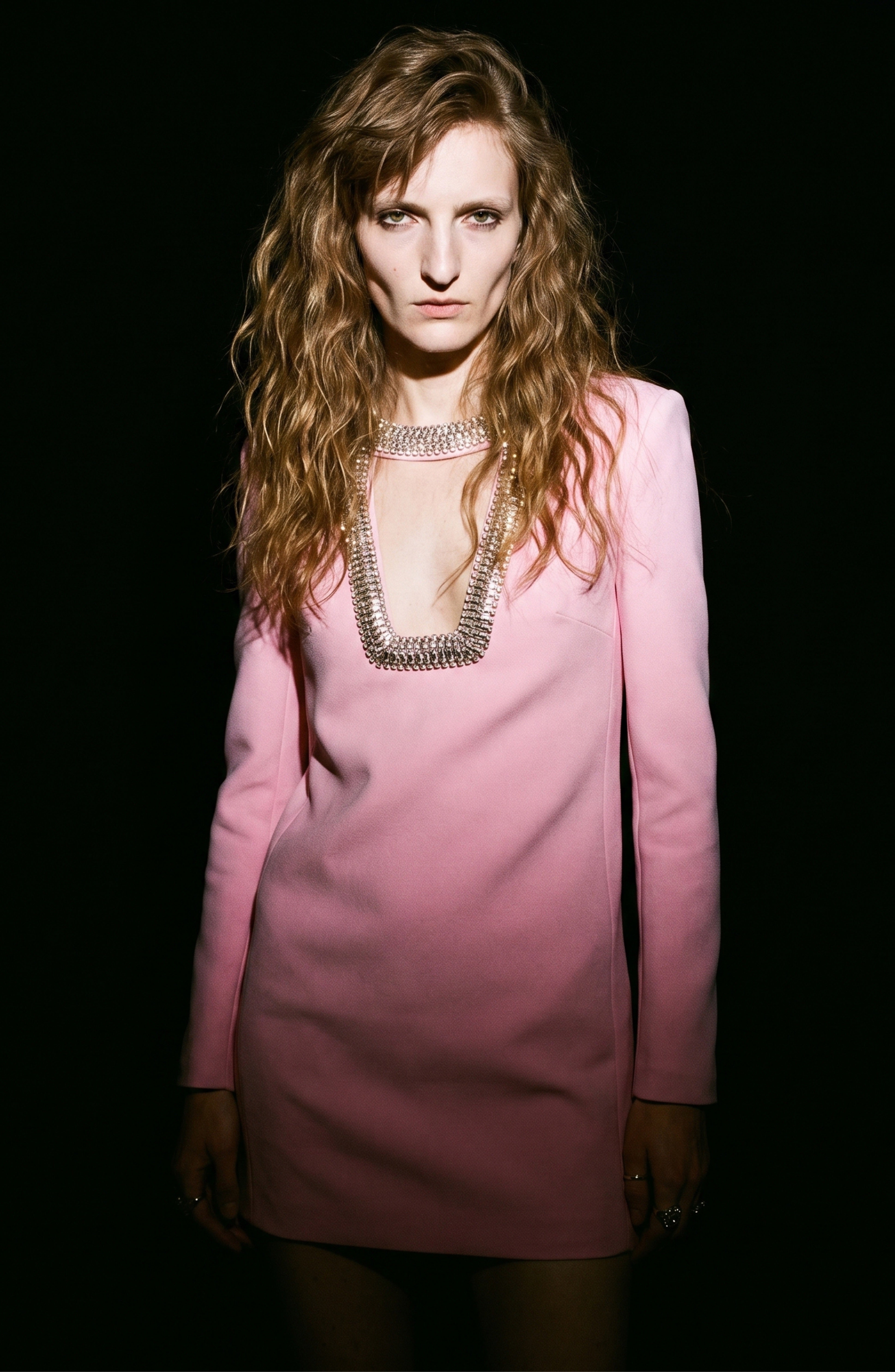 A person with long, wavy hair wears the LOULOU Crystal Cutout Mini Dress—a pastel pink mini dress featuring long sleeves, a deep neckline, and shimmering silver embellishments—standing against a black background.