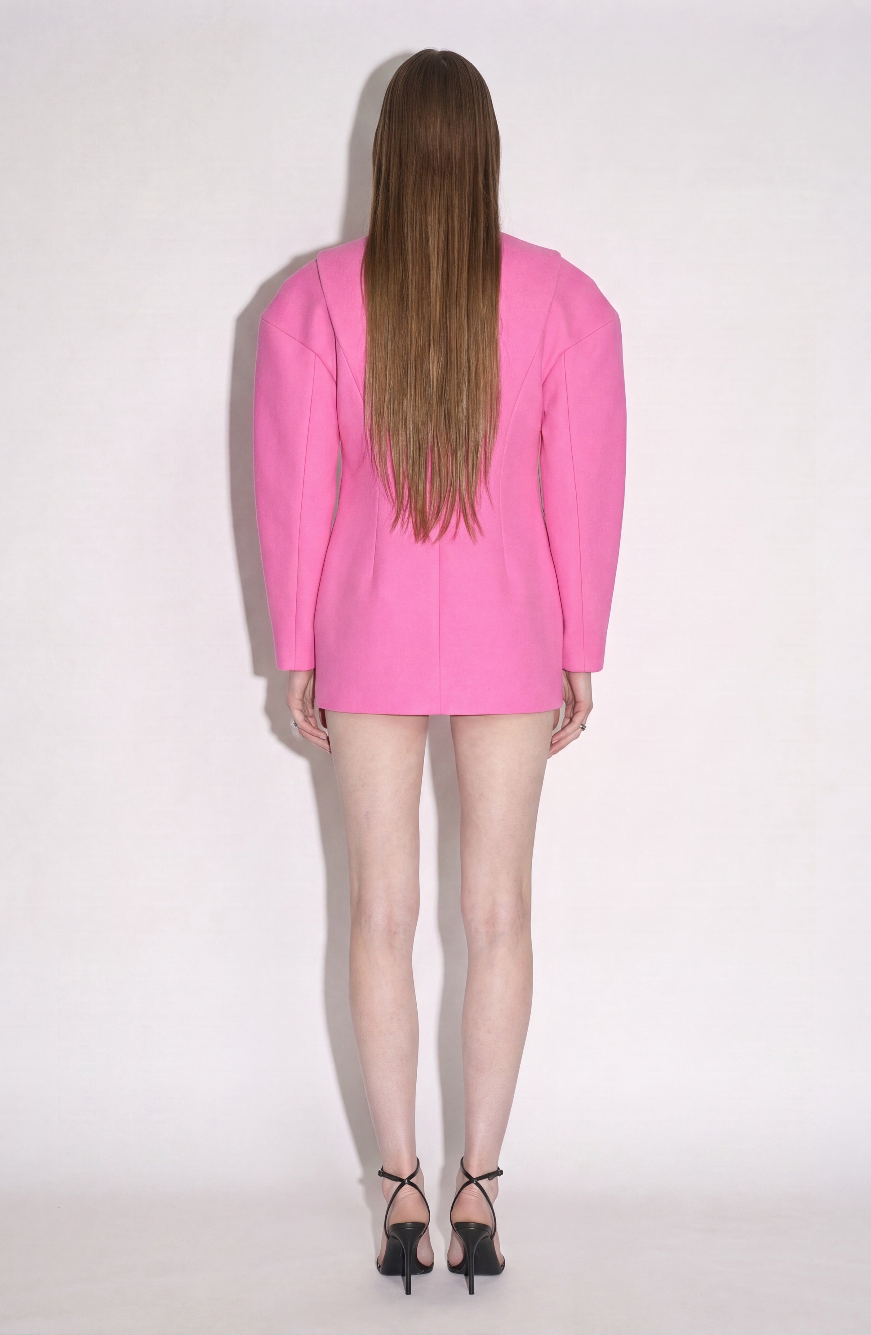 A person with long straight hair stands facing away, wearing the LOULOU Crystal Cutout Mini Dress in fuchsia, styled as a bright pink blazer, paired with black high-heeled sandals against a light, plain background.