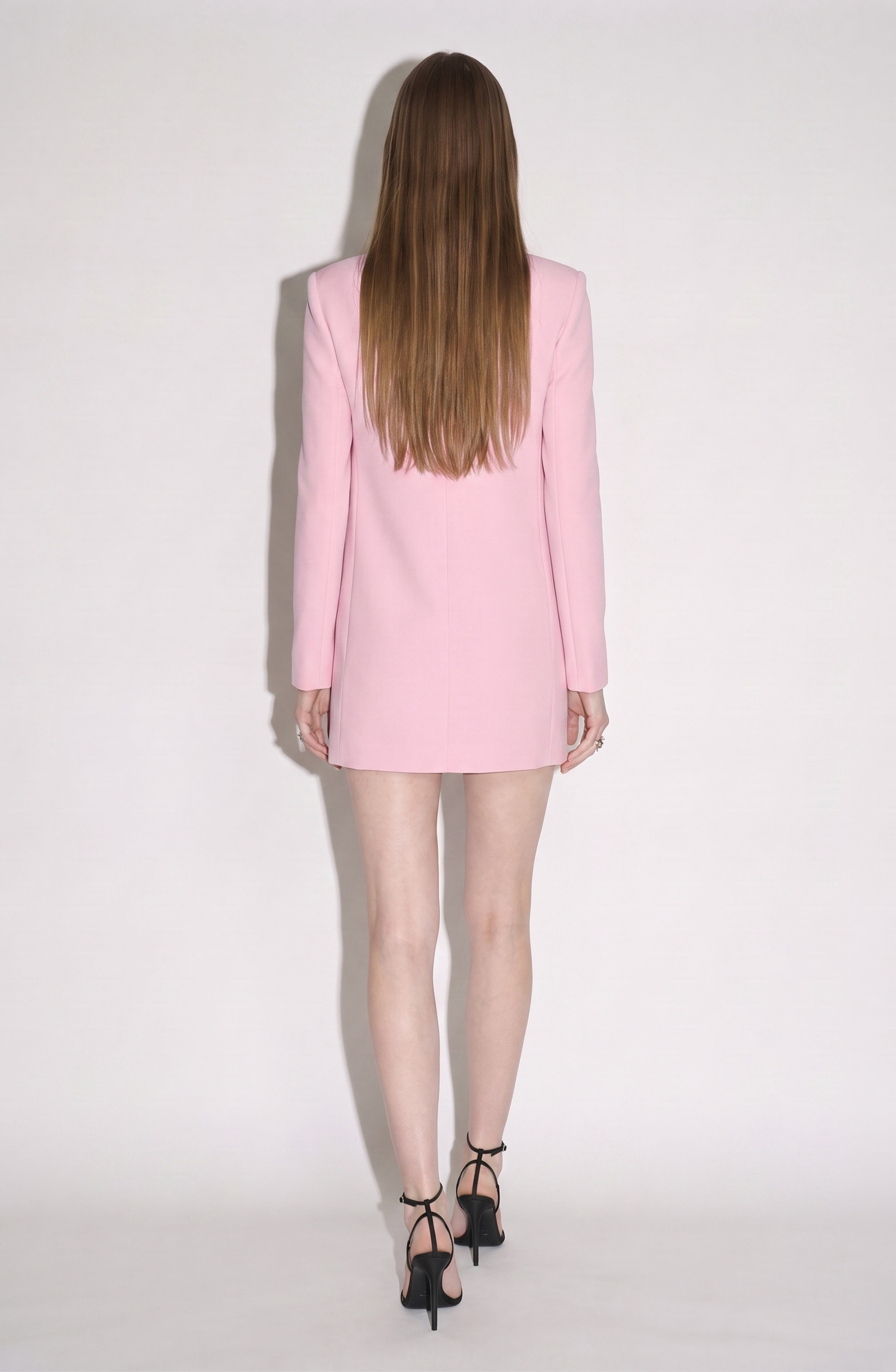 A woman with long straight hair stands facing away against a plain white background, wearing the LOULOU Crystal Cutout Mini Dress in pastel pink styled as a blazer, paired with black high-heeled sandals.