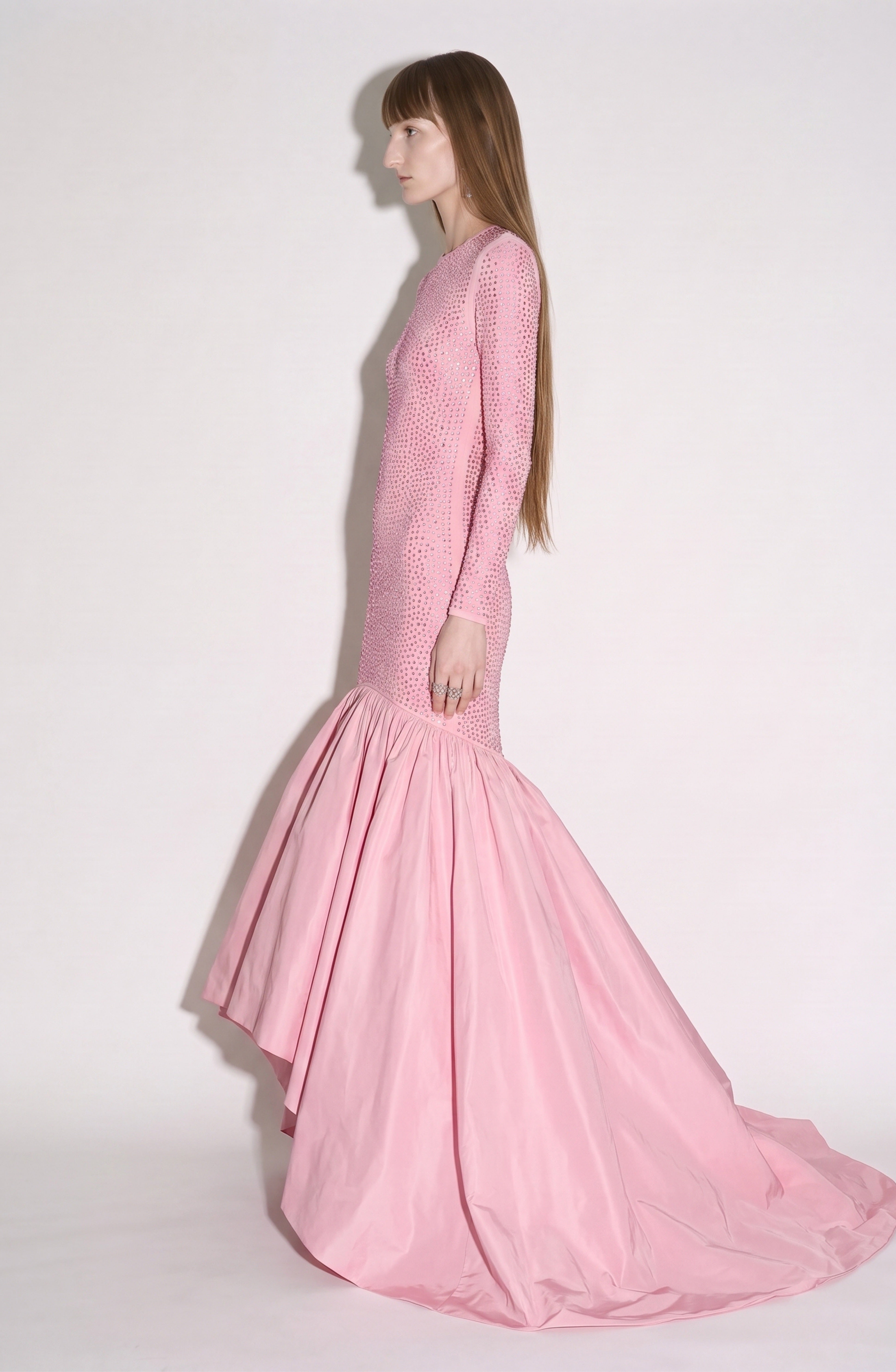 A person stands in profile wearing the LOULOU Crystal Embellished High-Low Gown, featuring long sleeves, a fitted silhouette with a crystal-embellished bodice, and a dramatic flared skirt. The plain white background accentuates the rose pink hue of the gown.