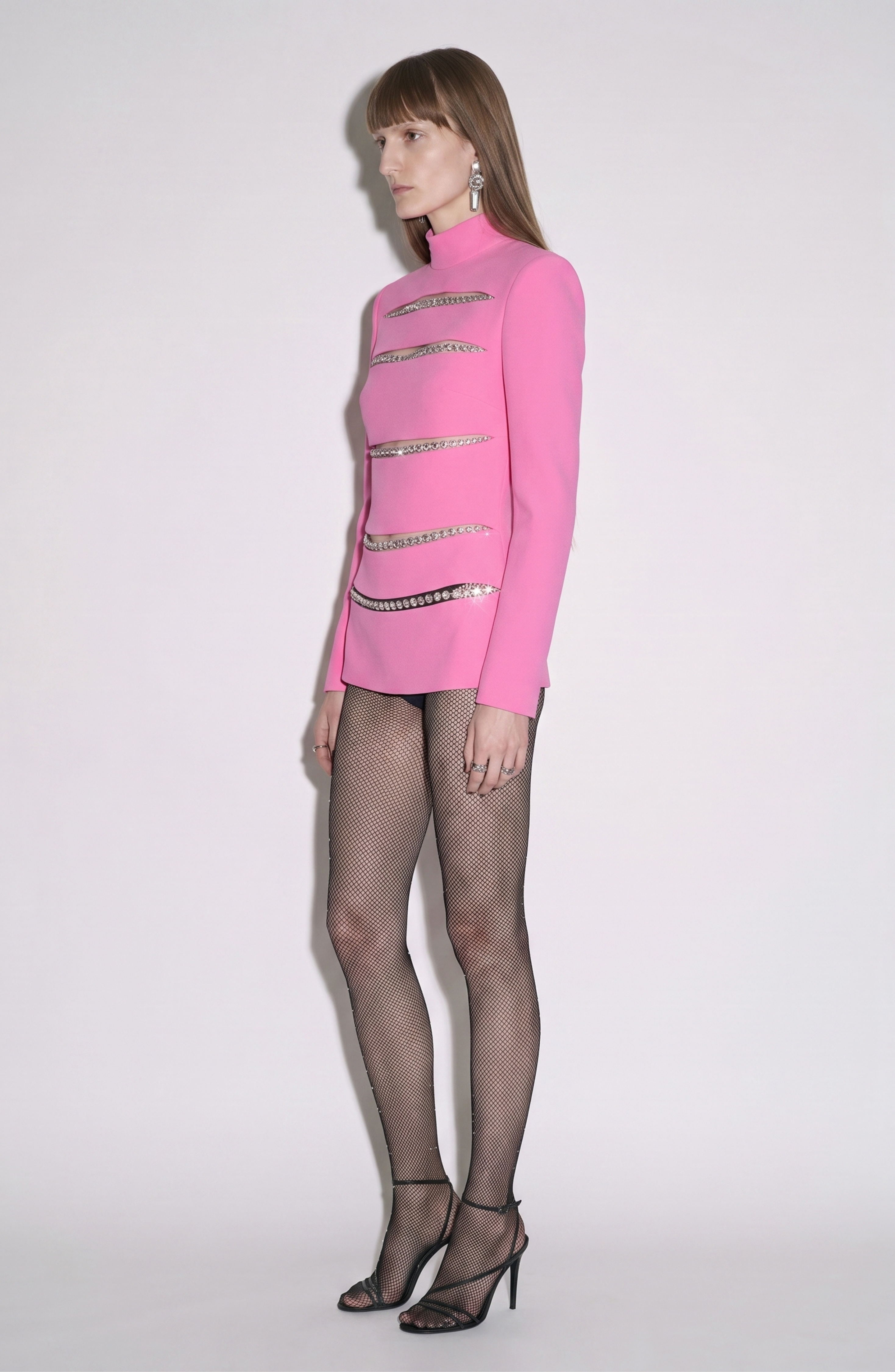 A person stands sideways wearing the LOULOU Crystal Slash Mini Dress against a plain white background, styled with fishnet tights and black high-heeled sandals.