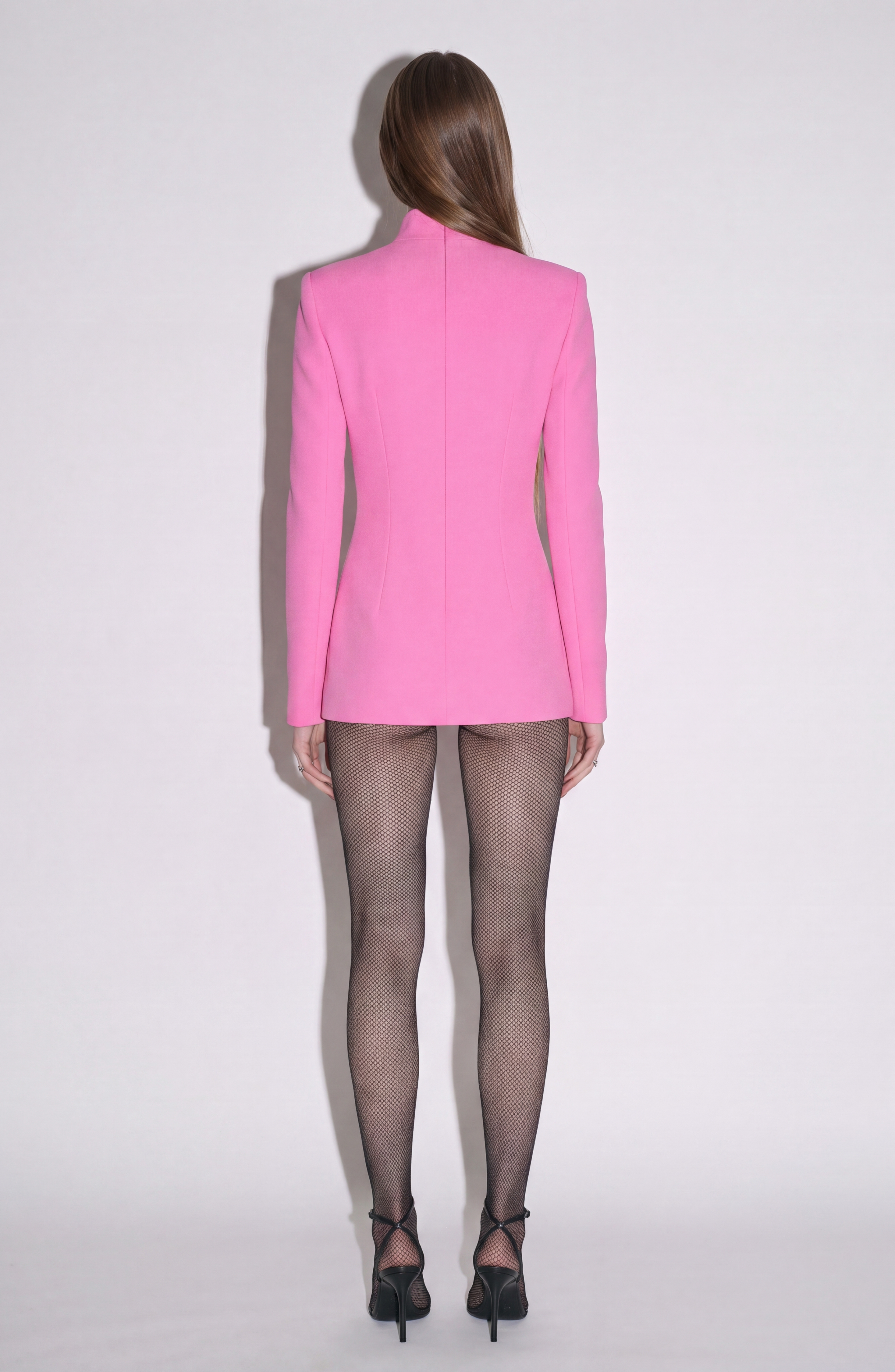 A person with long hair stands facing away, wearing the LOULOU Crystal Slash Mini Dress in fuchsia, paired with black fishnet tights and black high heels, against a plain light background.