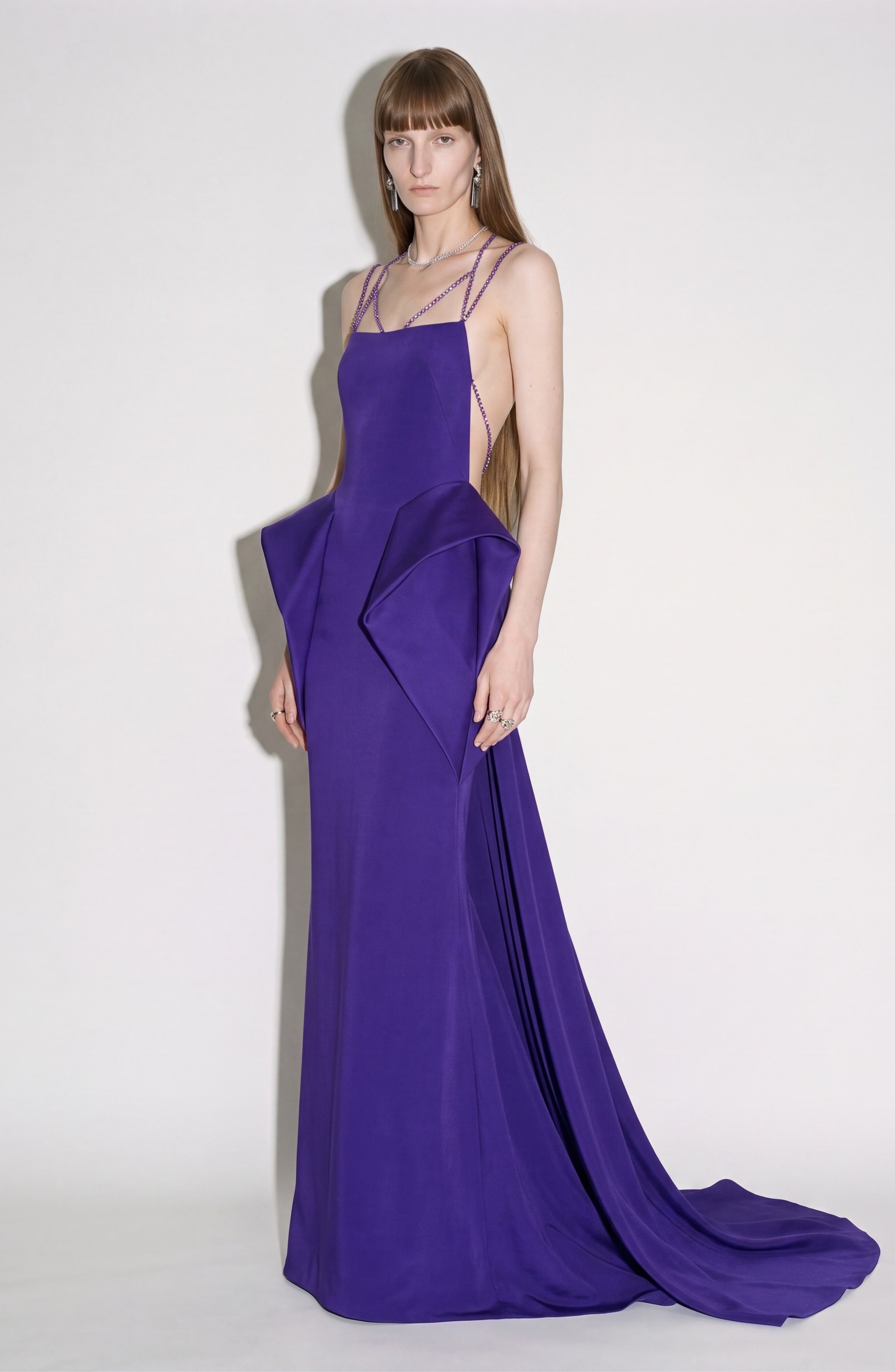 A person stands against a plain background, wearing the LOULOU Crystal Strap Peplum Gown—a floor-length, vibrant purple evening gown featuring geometric side panels, thin crystal chain straps, subtle structured peplums at the sides, and a small train.
