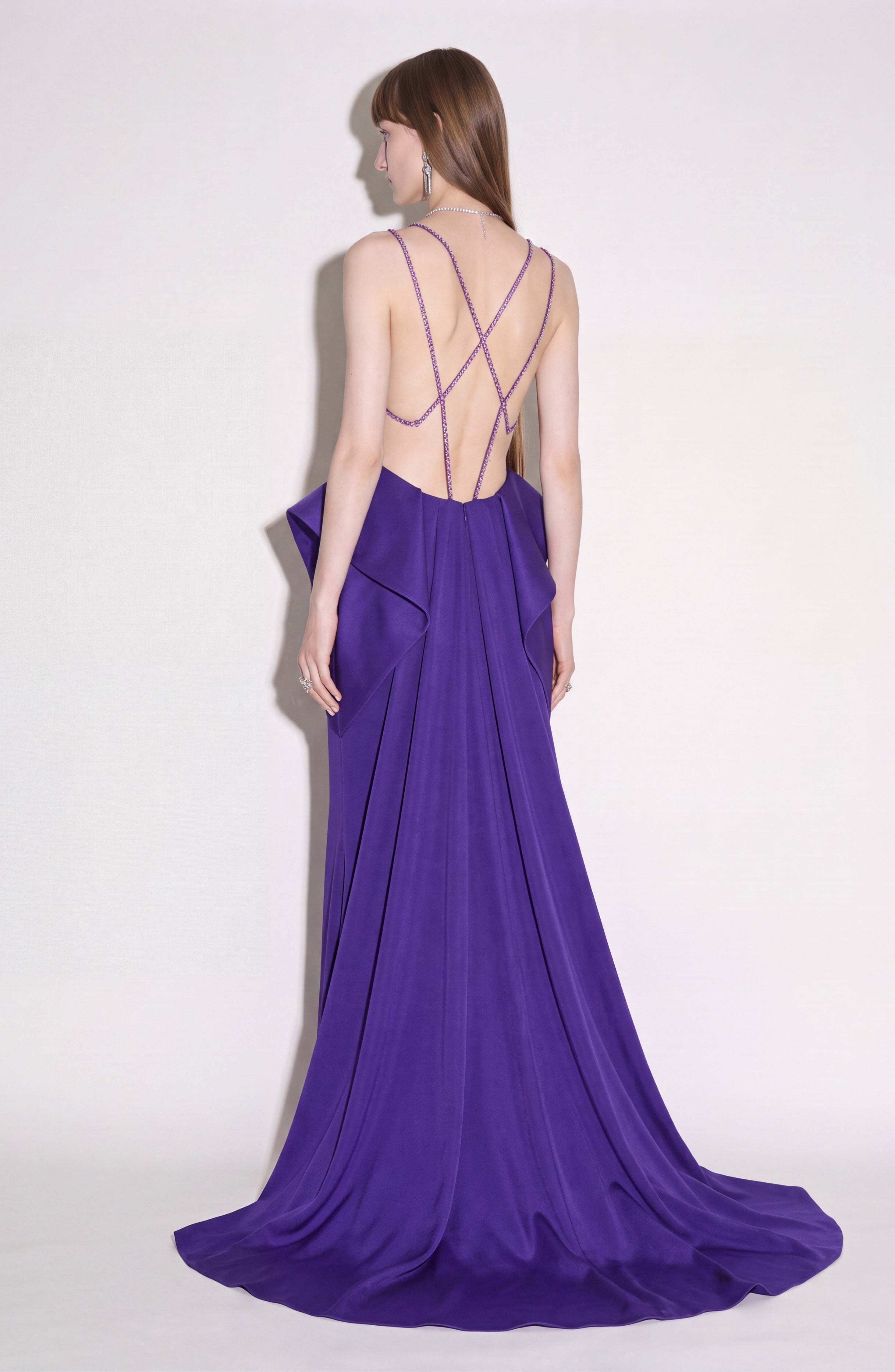 A woman with long straight hair models the LOULOU Crystal Strap Peplum Gown, a floor-length, backless purple evening dress featuring crystal chain straps and striking side panels, set against a plain light background.