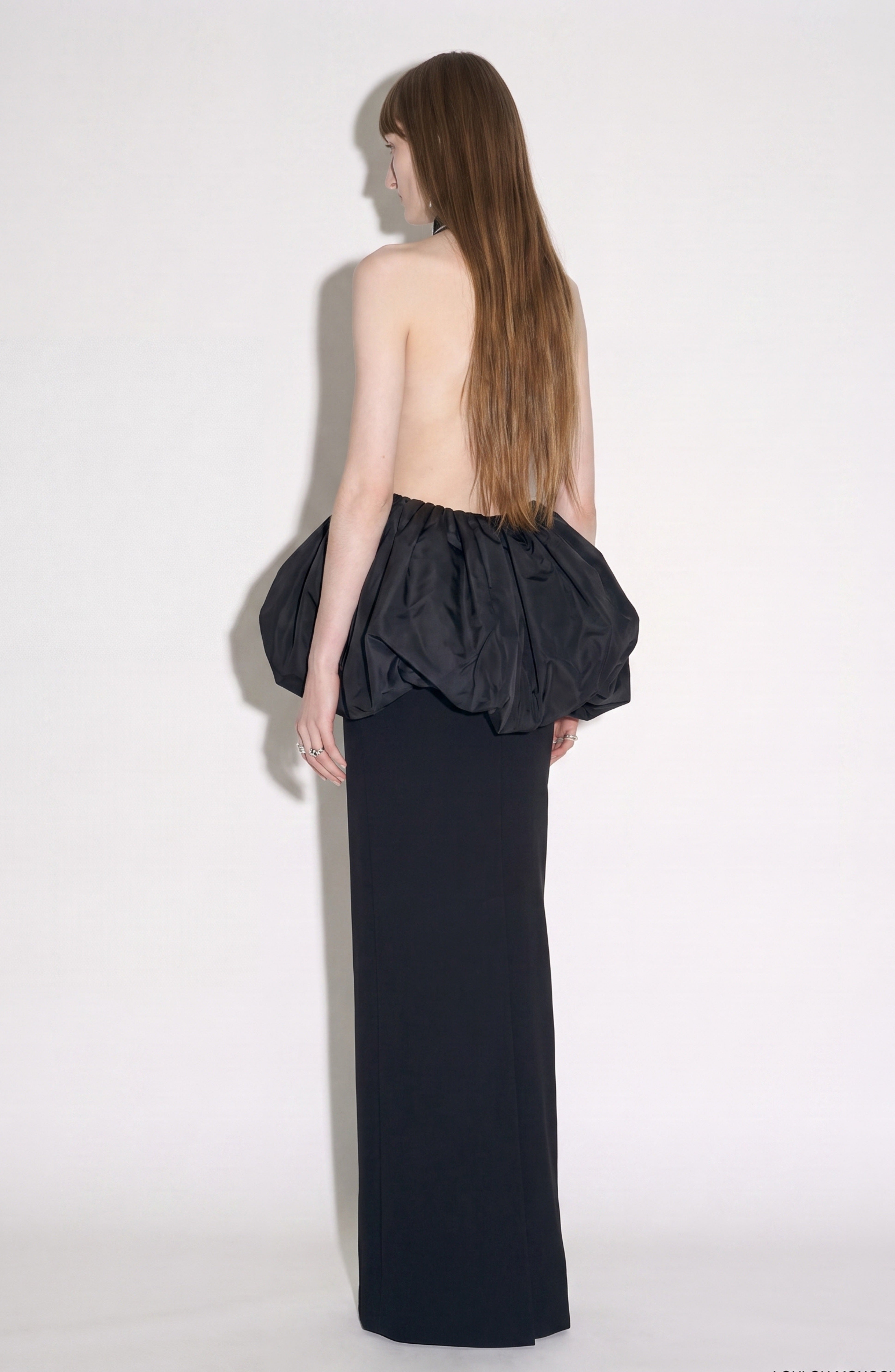 A person with long hair is seen from behind, standing against a plain light background and wearing the LOULOU Crystal Trim Peplum Maxi Dress, a backless black maxi dress with a voluminous skirt detail.