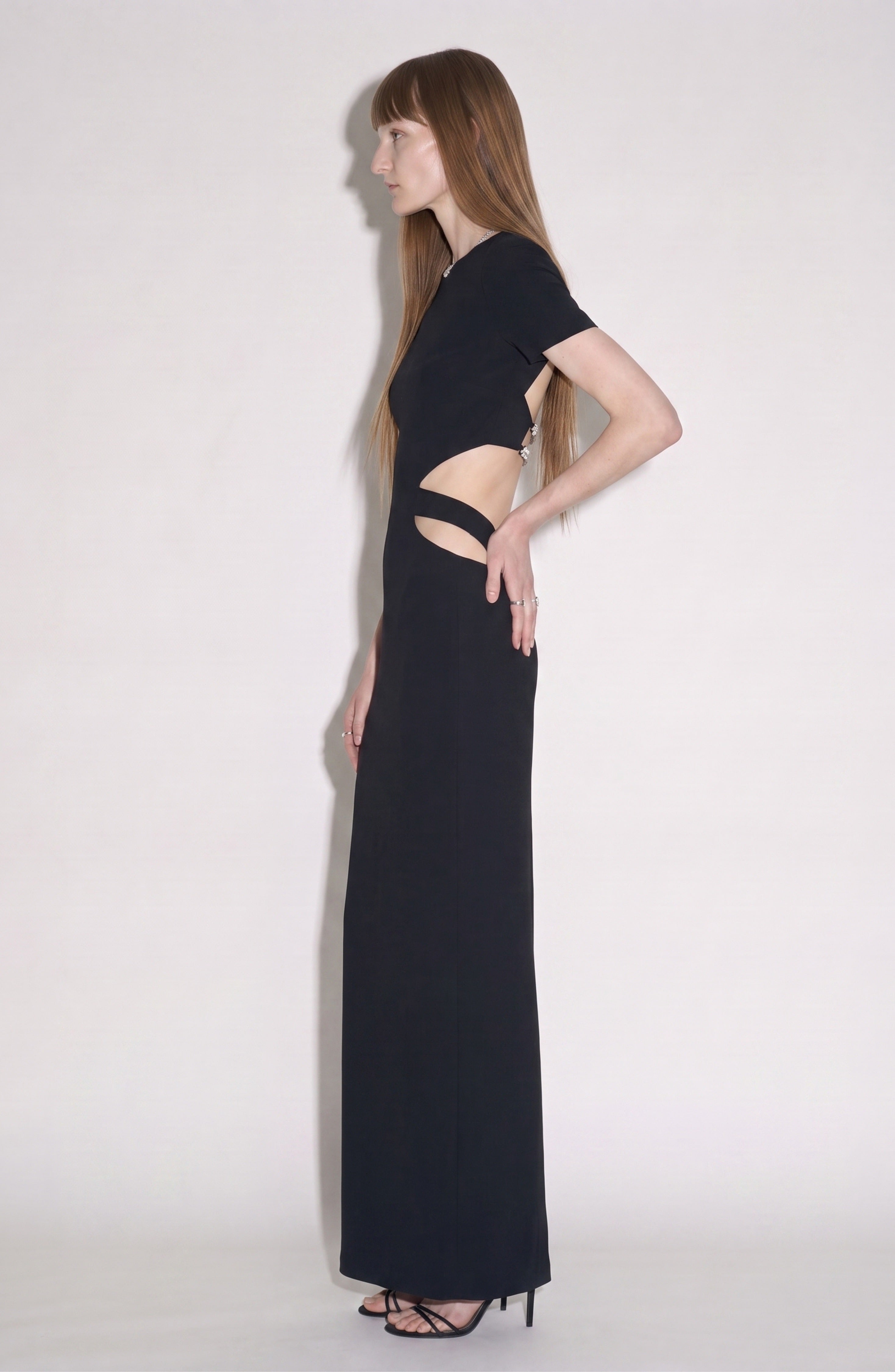 A woman stands in profile against a plain background wearing the LOULOU Cut-Out Maxi Dress in jet black, featuring waist cut-outs and short sleeves, paired with black high-heeled sandals adorned with crystal-embellished straps.