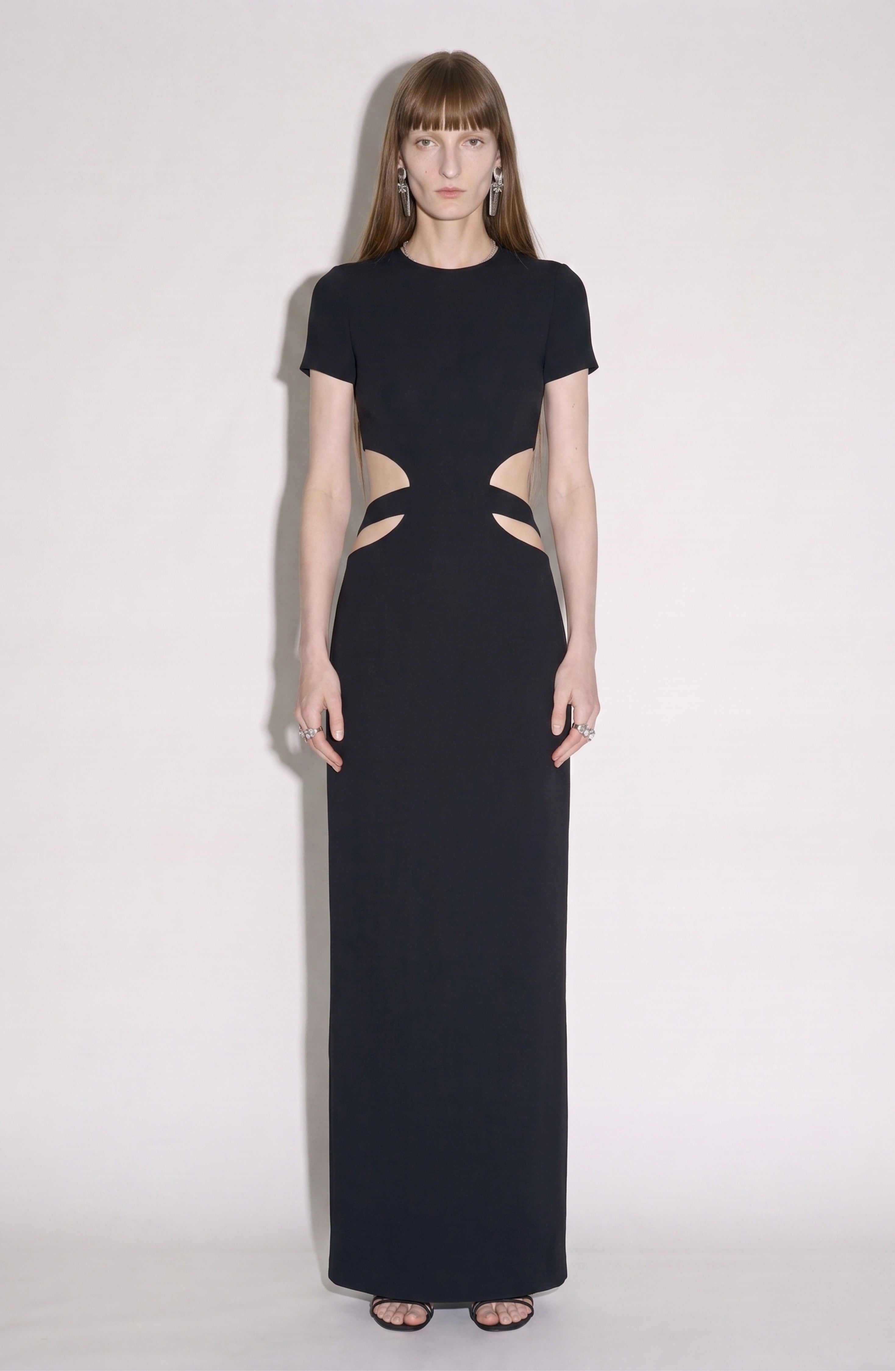 A woman wearing the LOULOU Cut-Out Maxi Dress in jet black, featuring short sleeves and waist cut-outs, stands against a plain white background, paired with black sandals and minimal jewelry.