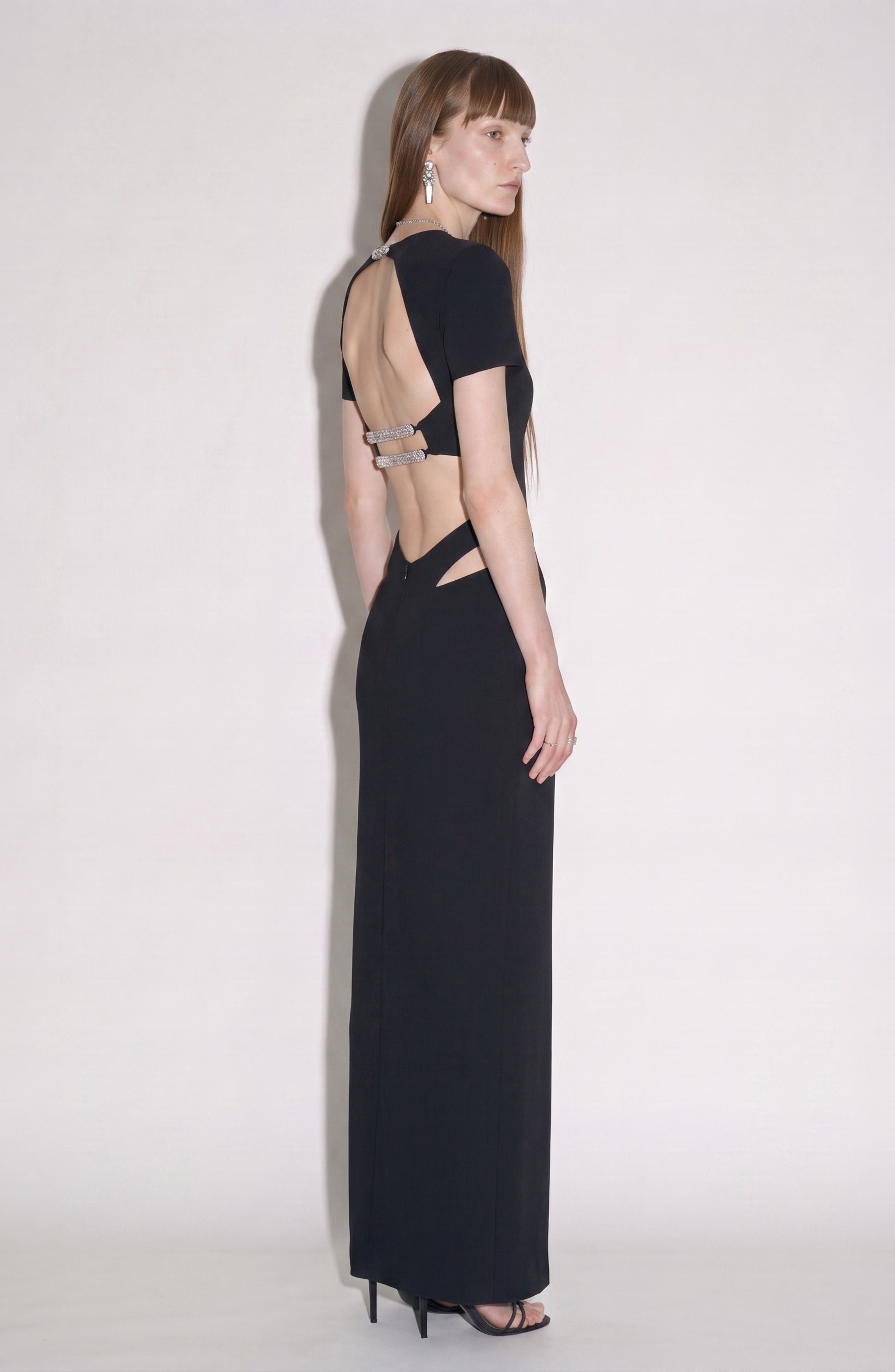 A woman in LOULOU’s Cut-Out Maxi Dress—featuring a jet black hue, crystal-embellished straps, and an open back—stands sideways, glancing over her shoulder. She pairs the look with black heels and wears her long, straight brown hair down.
