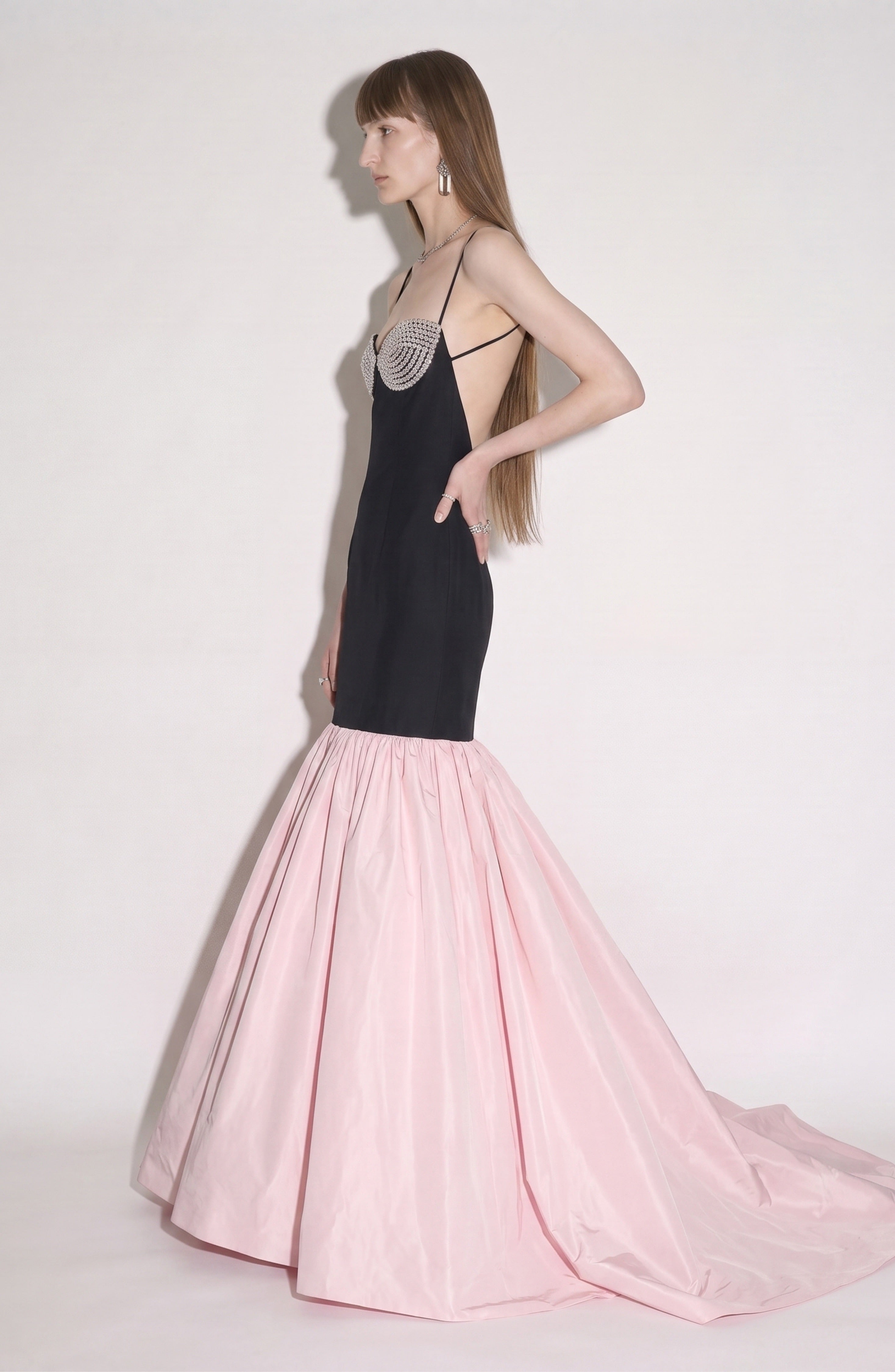 A woman stands in profile against a plain background, wearing the LOULOU Detailed Bustier Mermaid Gown—a sleeveless black and pink dress featuring a fitted bodice with crystal-embellished bust detail and a flared, floor-length taffeta skirt.