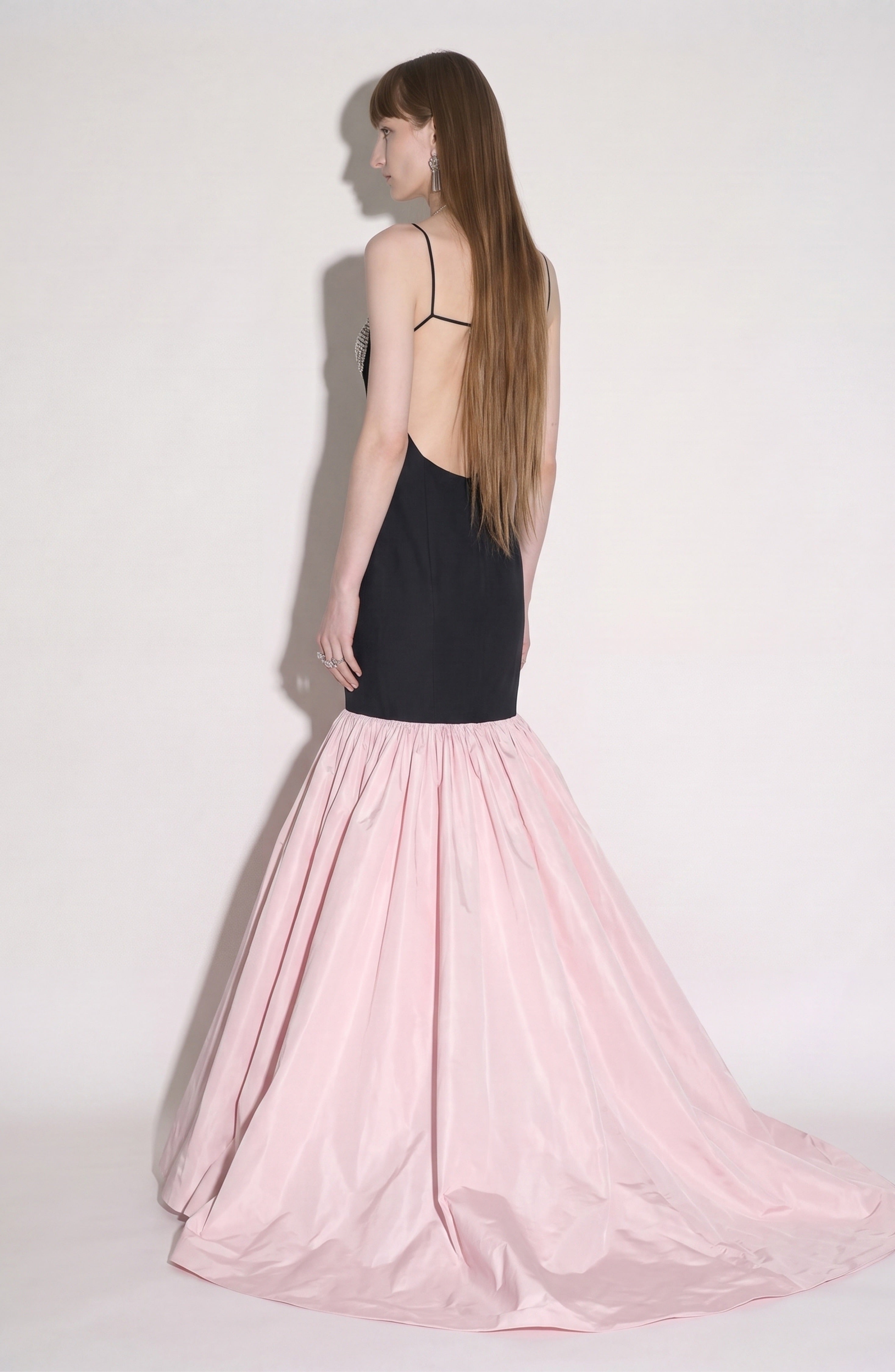 A woman with long, straight hair stands in profile against a plain background, wearing the LOULOU Detailed Bustier Mermaid Gown—a black design with thin straps and a dramatic, crystal-embellished flared light pink taffeta skirt.