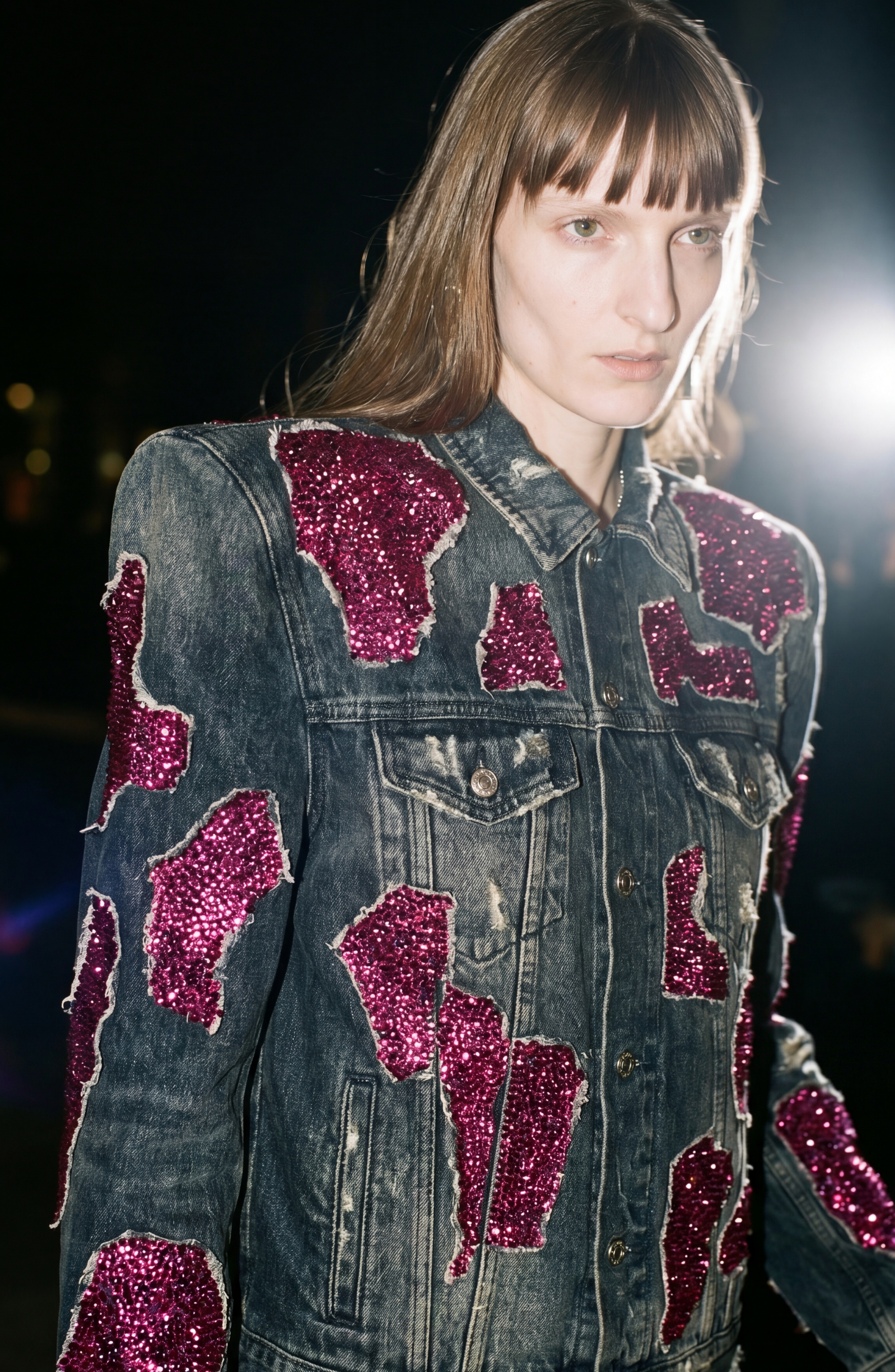 A person wearing the LOULOU Embellished Denim Jacket, featuring a vintage-washed finish and striking magenta sequin appliqués, stands in dramatic lighting with wet-look hair and a serious expression.